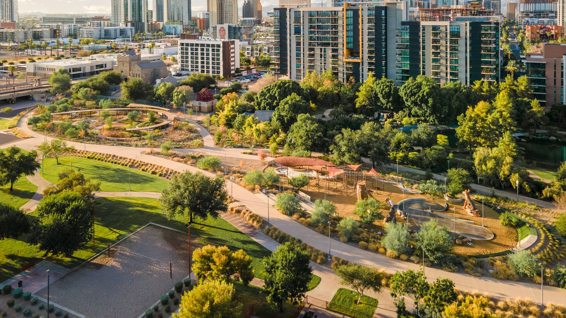 Aerial cityscape with high-rise glass buildings in the background, a lush urban park with winding paths, trees, and playgrounds in the foreground, under a blue sky with scattered clouds.