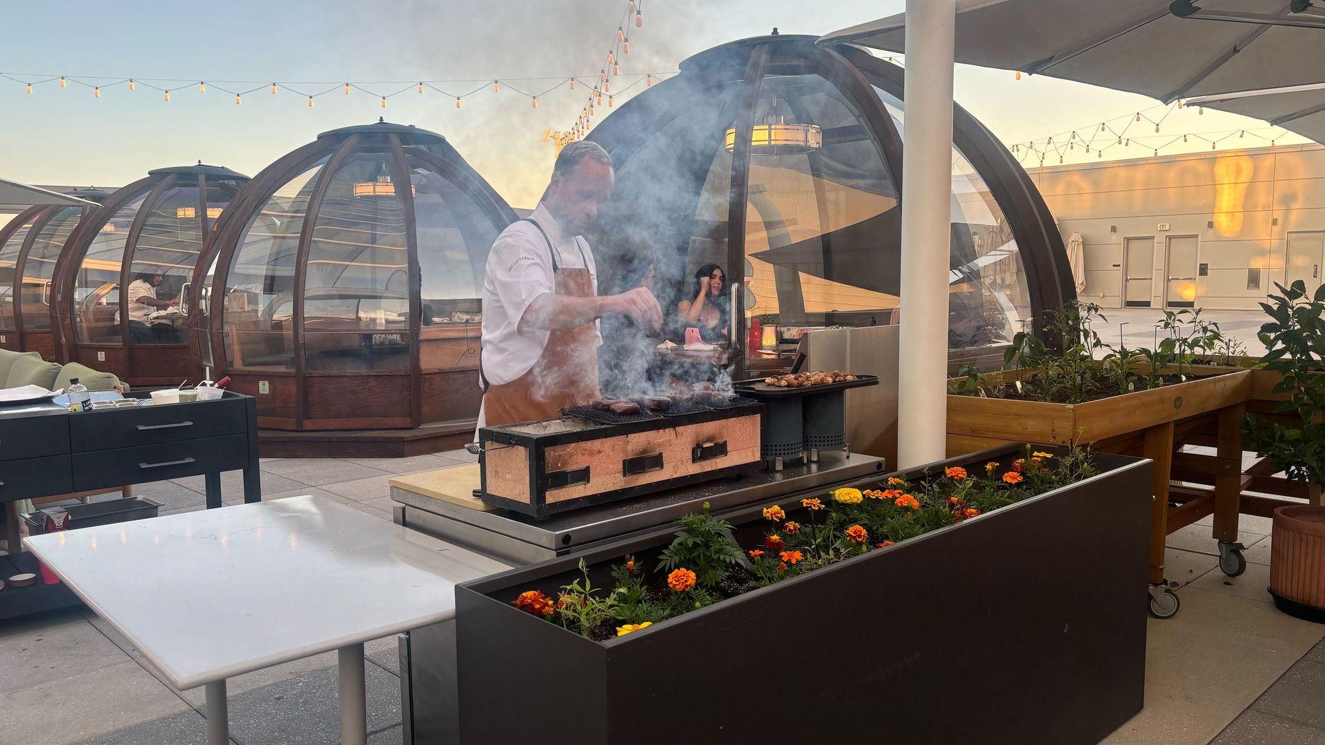 Chef in white shirt and brown apron grills on a rooftop BBQ as smoke rises; glass-domed grills, string lights, orange flowering planters, and a person seated in the background.