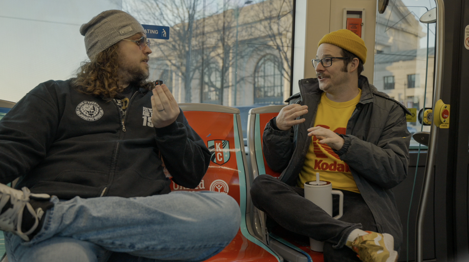 Two men sitting face-to-face on red and blue bus seats, one wearing a grey beanie and black jacket, the other in a yellow beanie and Kodak shirt, engaged in conversation.
