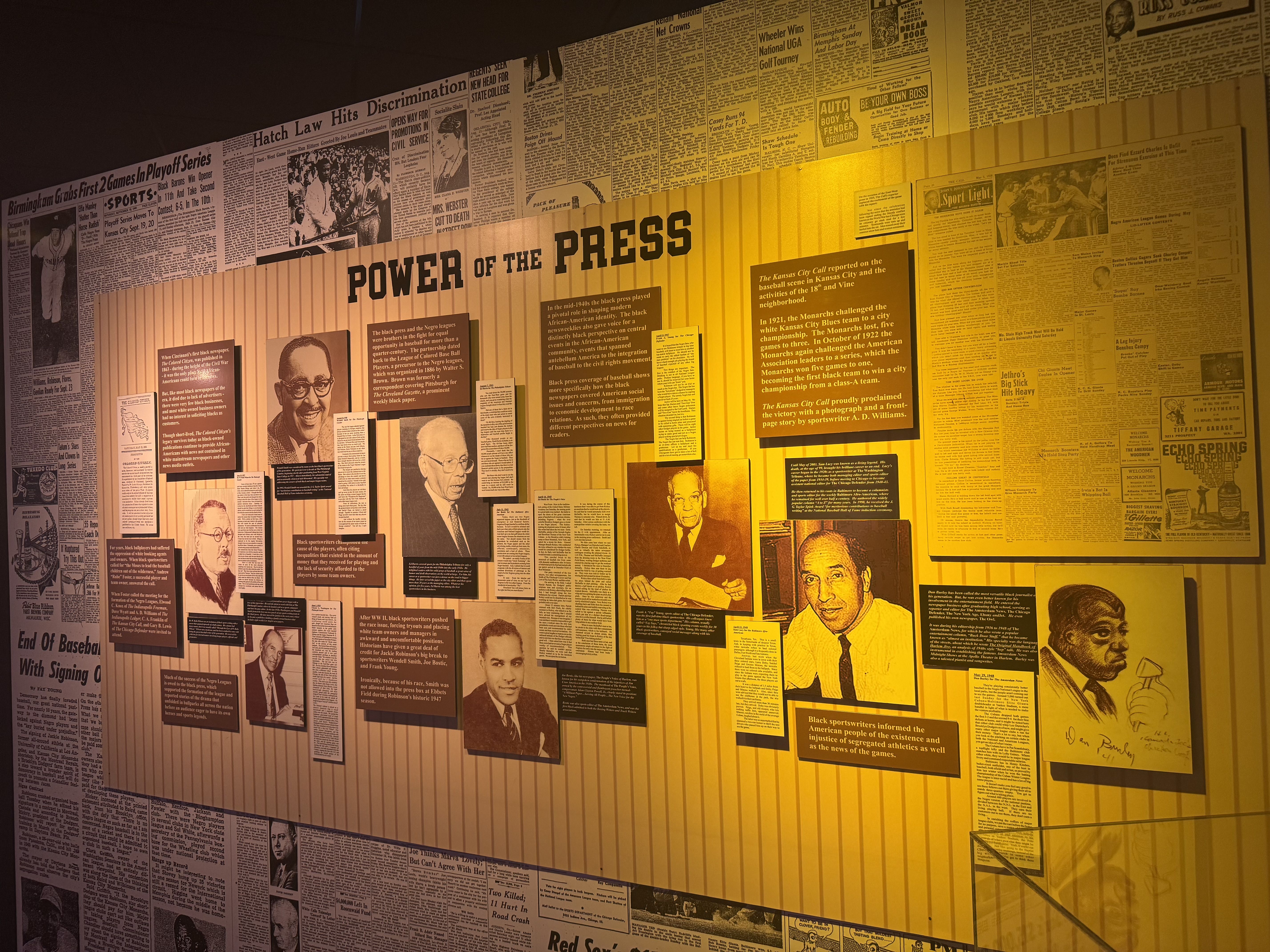 Exhibit wall with vintage newspaper clippings and photos arranged around a yellow panel reading "POWER OF THE PRESS", featuring portraits and captions under warm lighting.