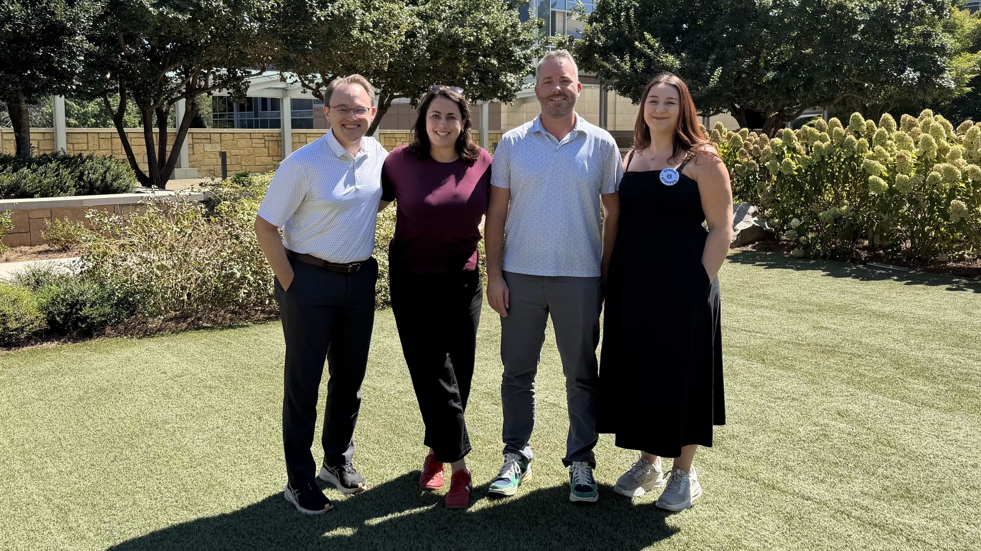 Four adults pose outdoors on a sunny day in front of glass office towers and greenery. From left: man in a light polo, woman in maroon, man in a light shirt, woman in a black dress with a badge.