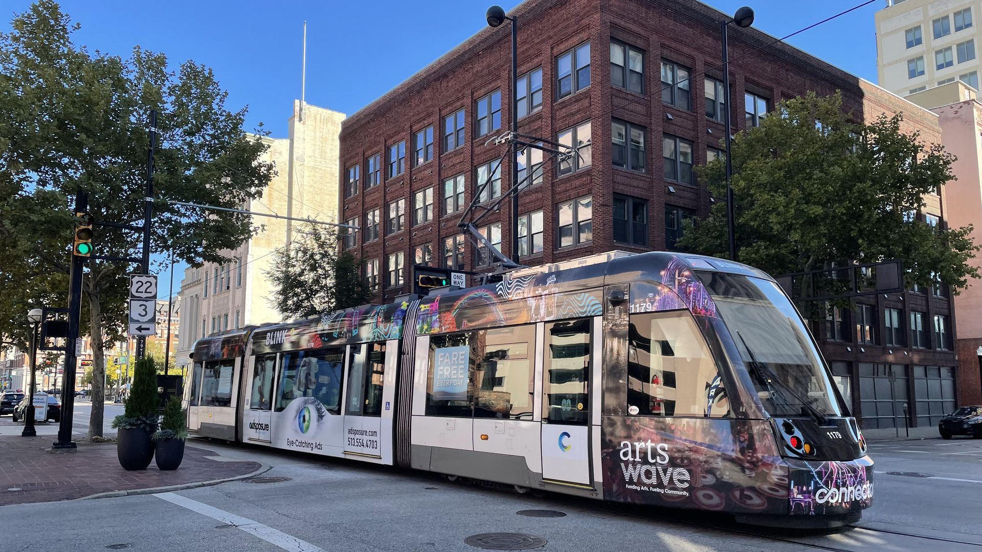 Colorful graffiti tram on a city street beside a red brick building under a bright blue sky, with green trees and traffic lights; a beige high-rise rises in the background.