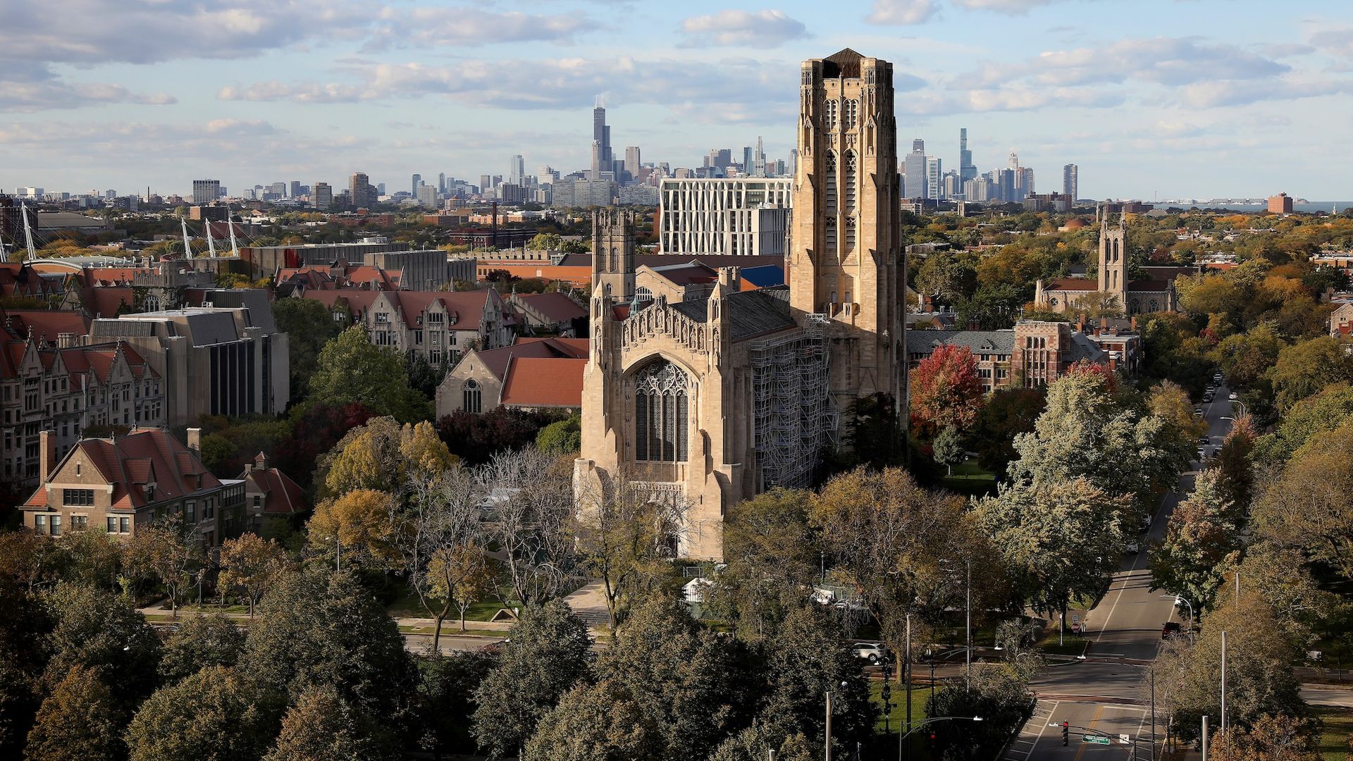 Wide view of a university campus with a tall Gothic stone tower in the foreground, red-roofed buildings and trees, and Chicago's downtown skyline in the distance under a blue, cloudy sky.