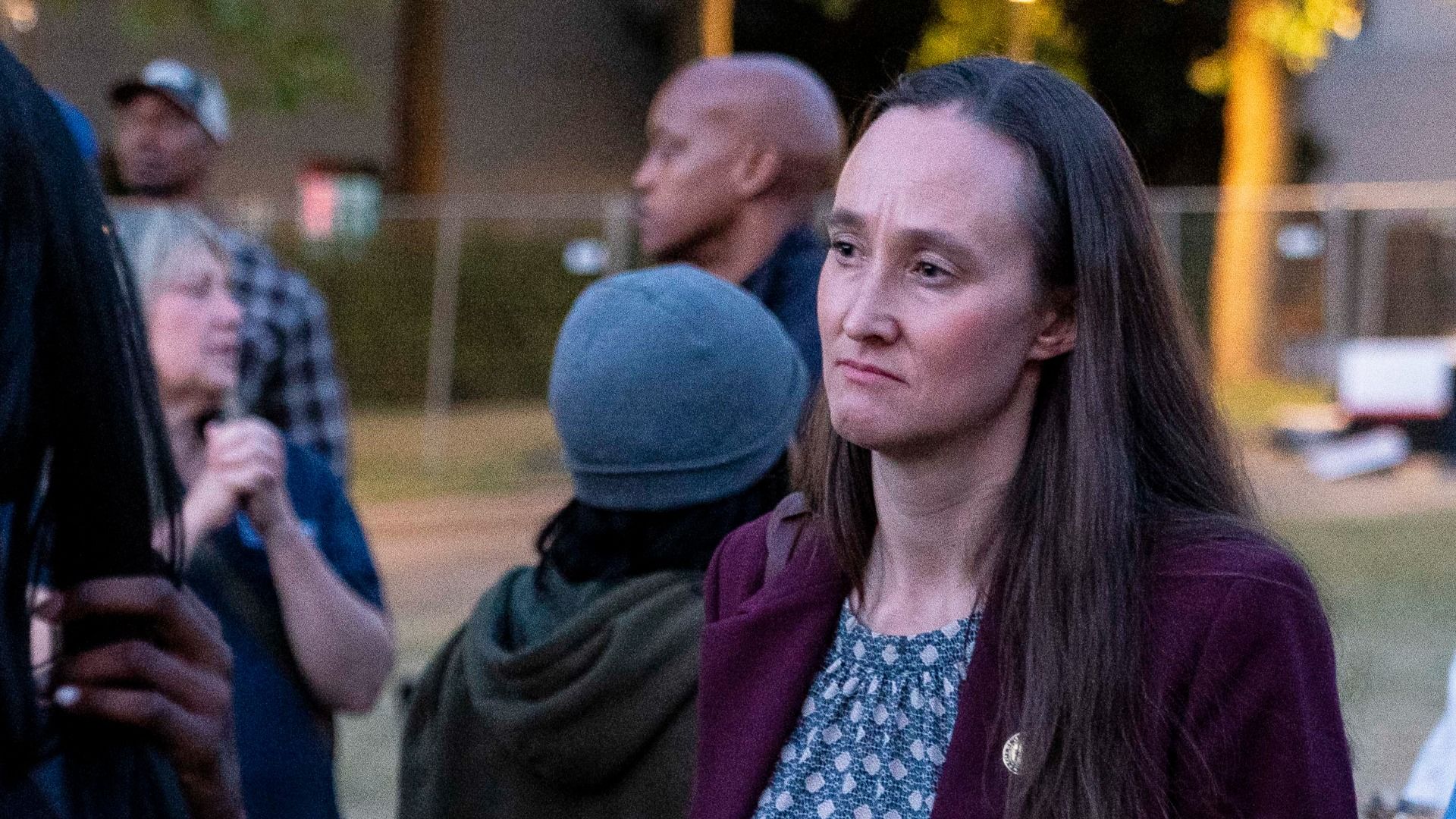 A woman with long brown hair wearing a maroon blazer over a blue patterned top stands outdoors at dusk, with blurred people and a fence in the background.