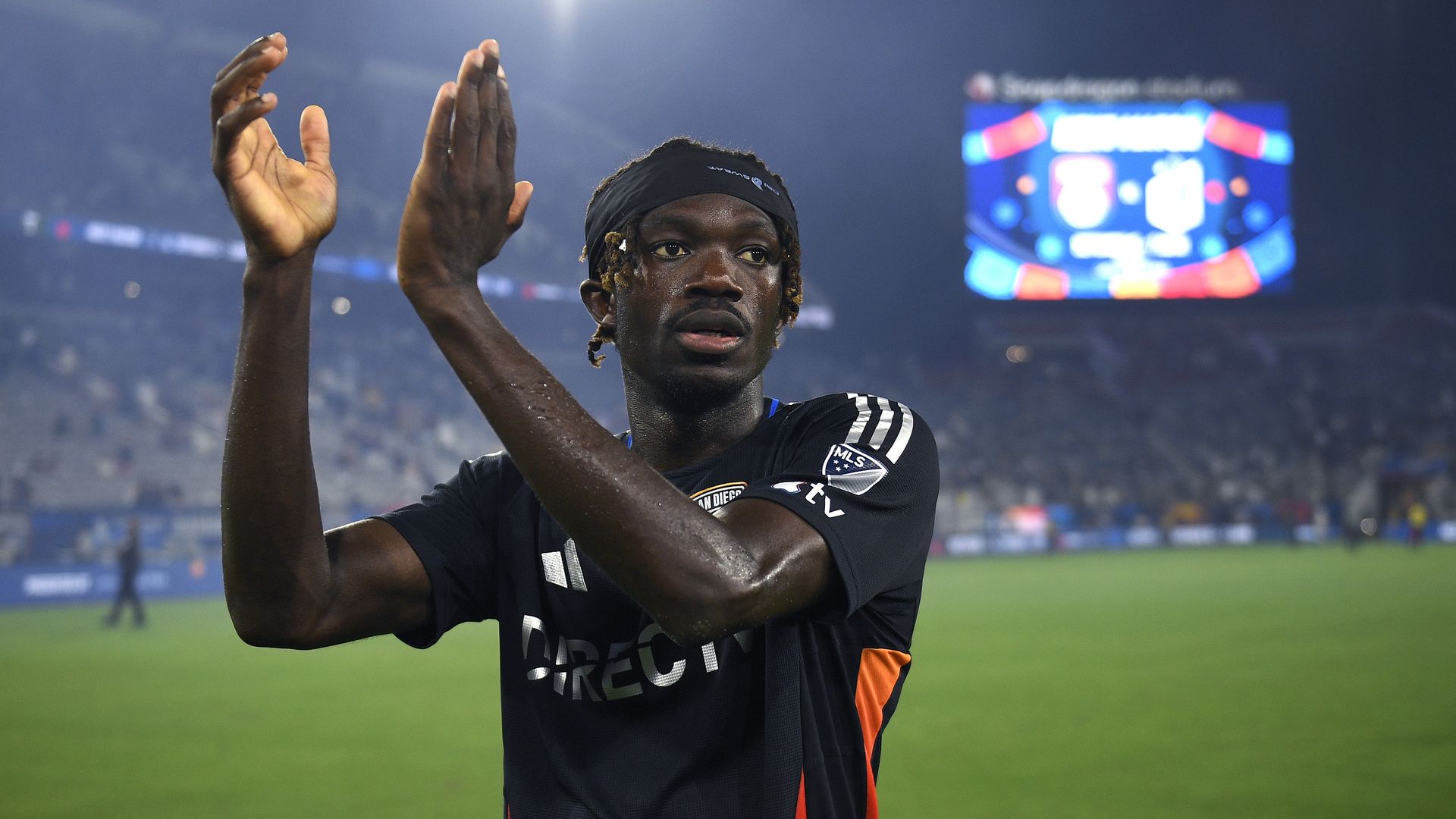 A soccer player in a black and orange jersey claps his hands on the field, with a blurred crowd and scoreboard visible in the background during a night game.