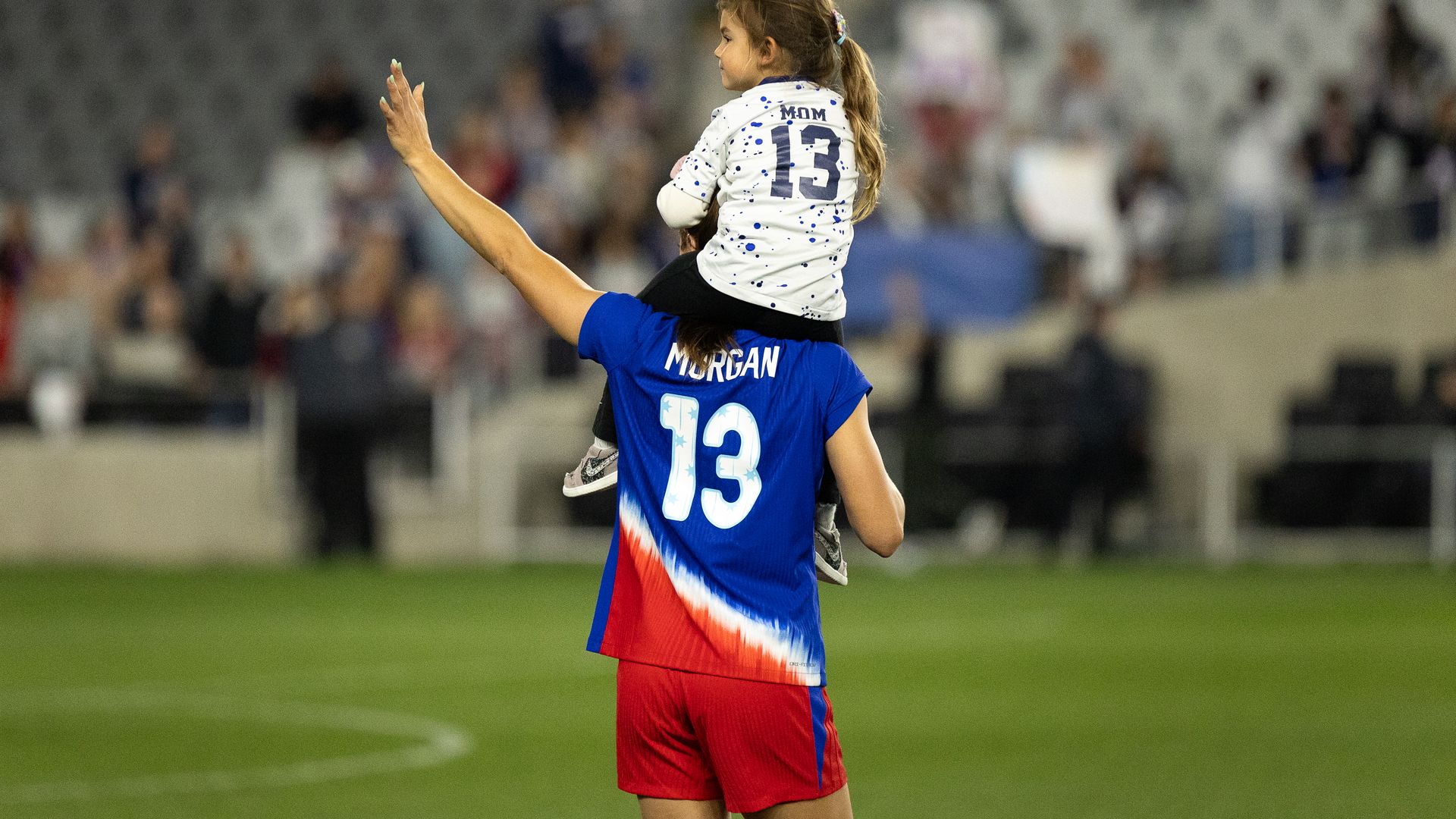 Alex Morgan waives to fans with her daughter on her shoulders