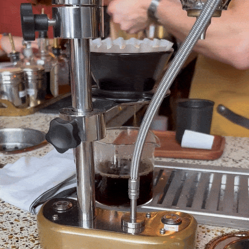 Coffee dripping into a glass beaker while baristas in yellow aprons work in the background.