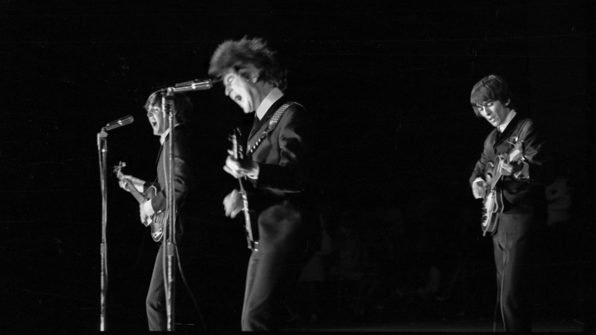 Black-and-white photo of a three-person rock band performing on stage: singer with mic on the left, two guitarists in the center and right, dark background and vintage concert vibe.