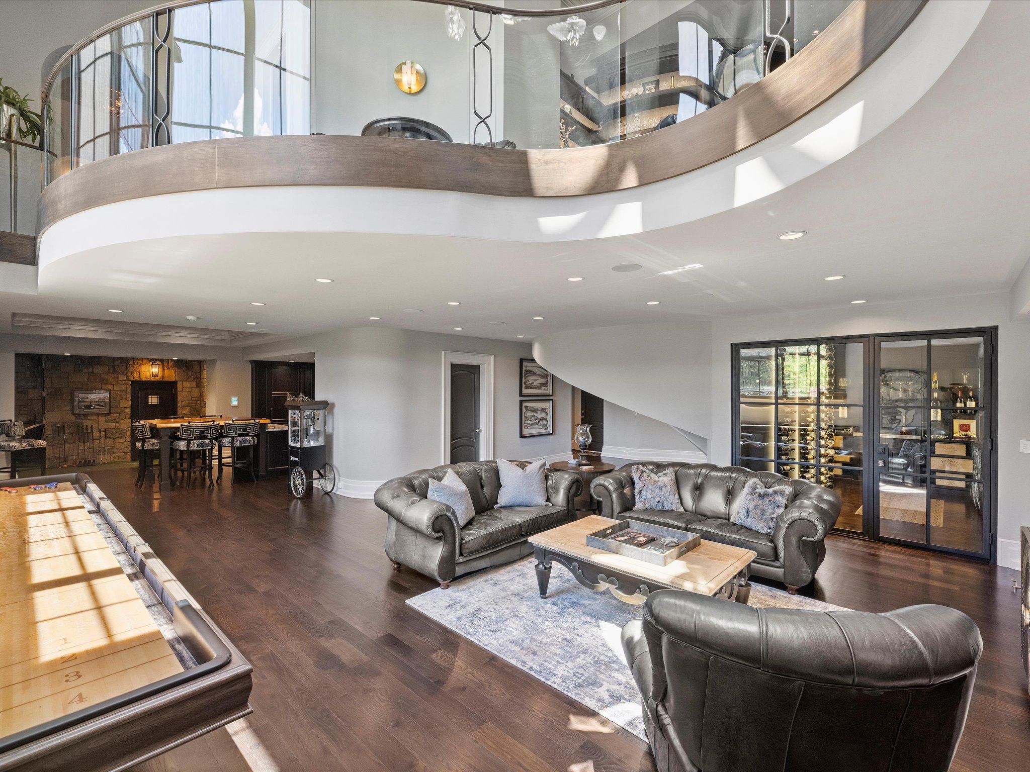 Spacious modern living area with dark leather sofas, a wooden coffee table on a gray rug, glass wine cellar, a bar with stools, and a shuffleboard table on dark wood flooring.