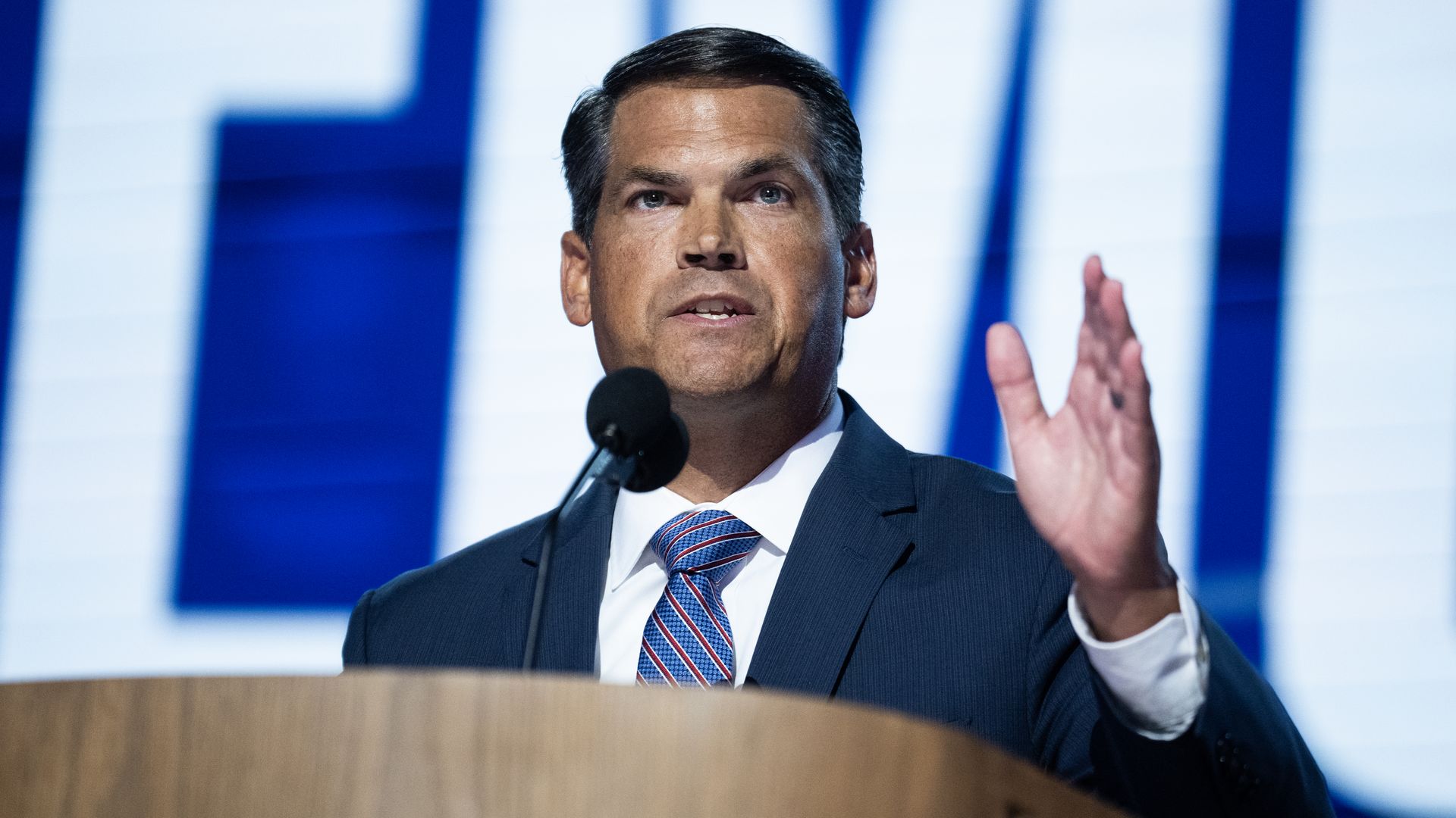 A man wearing a dark-colored suit, a white collared shirt and blue tie with red and white stripes holds his left hand in the air as he speaks into a microphone protruding from a light brown podium.