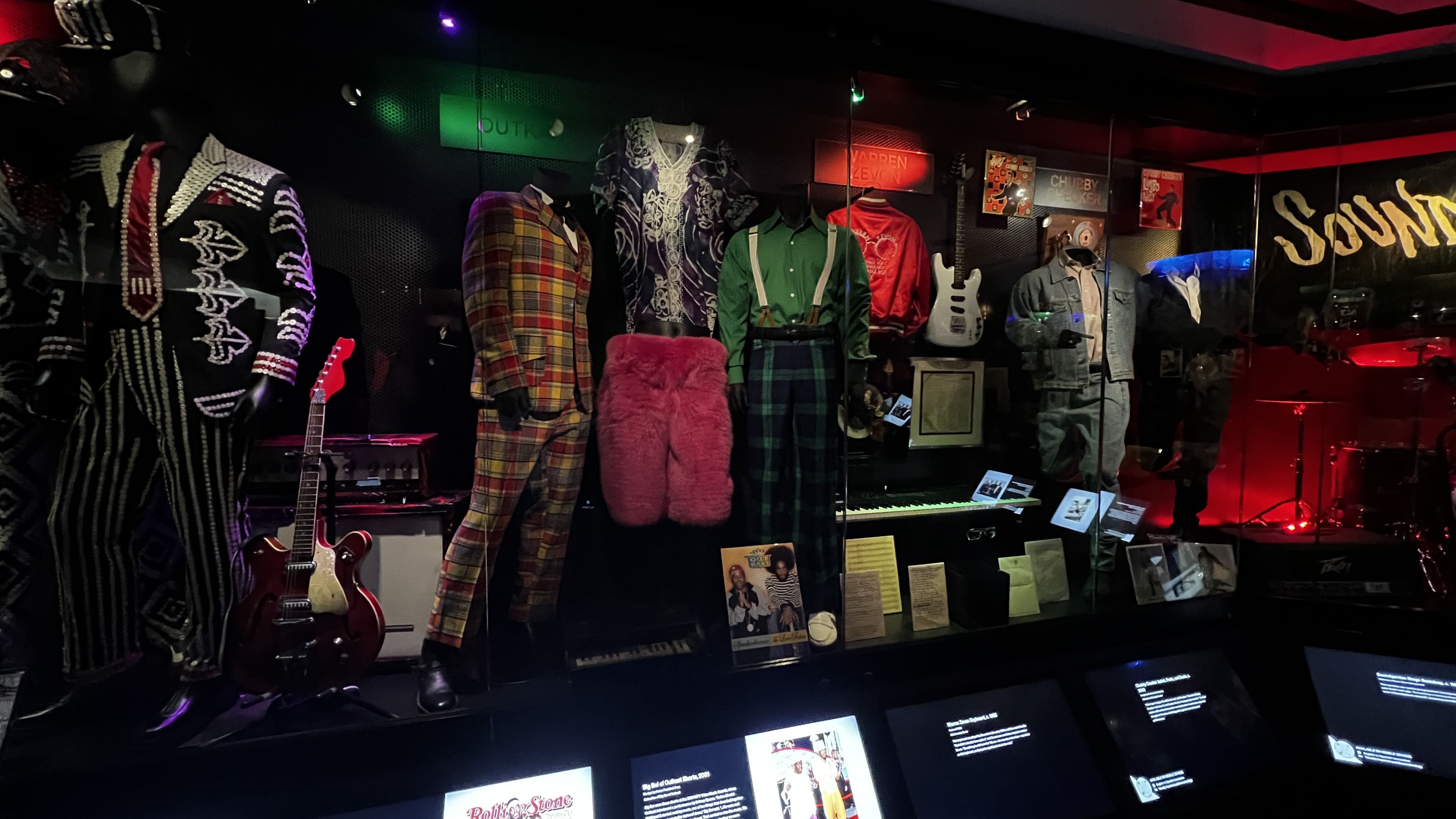 Display of colorful vintage band costumes including striped suits, pink furry pants, and plaid outfits behind glass with guitars and memorabilia in a dimly lit music exhibit.