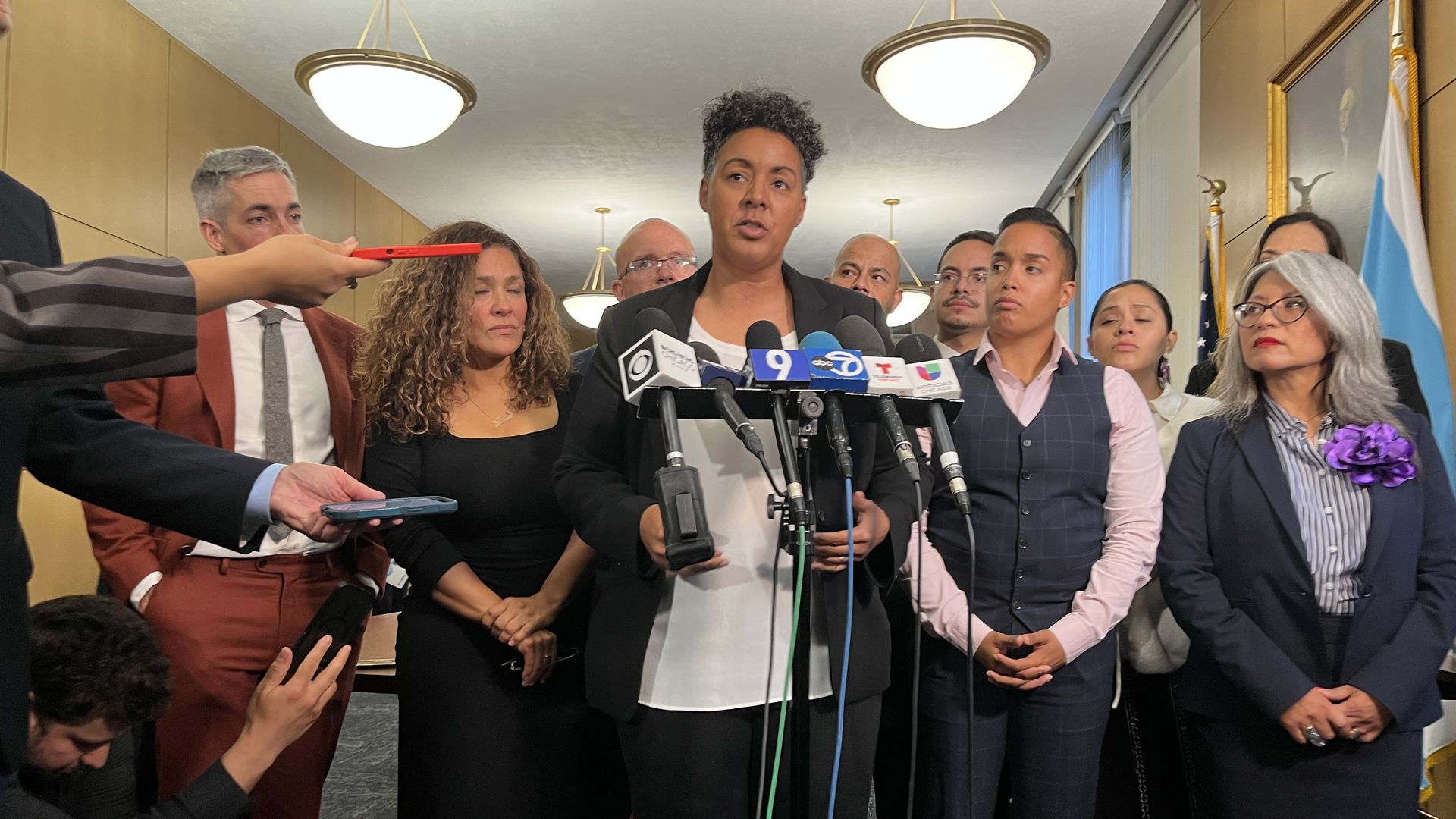 Woman in black suit and white top speaking at microphones during a press conference, surrounded by people in formal attire, some holding phones and recording devices indoors.