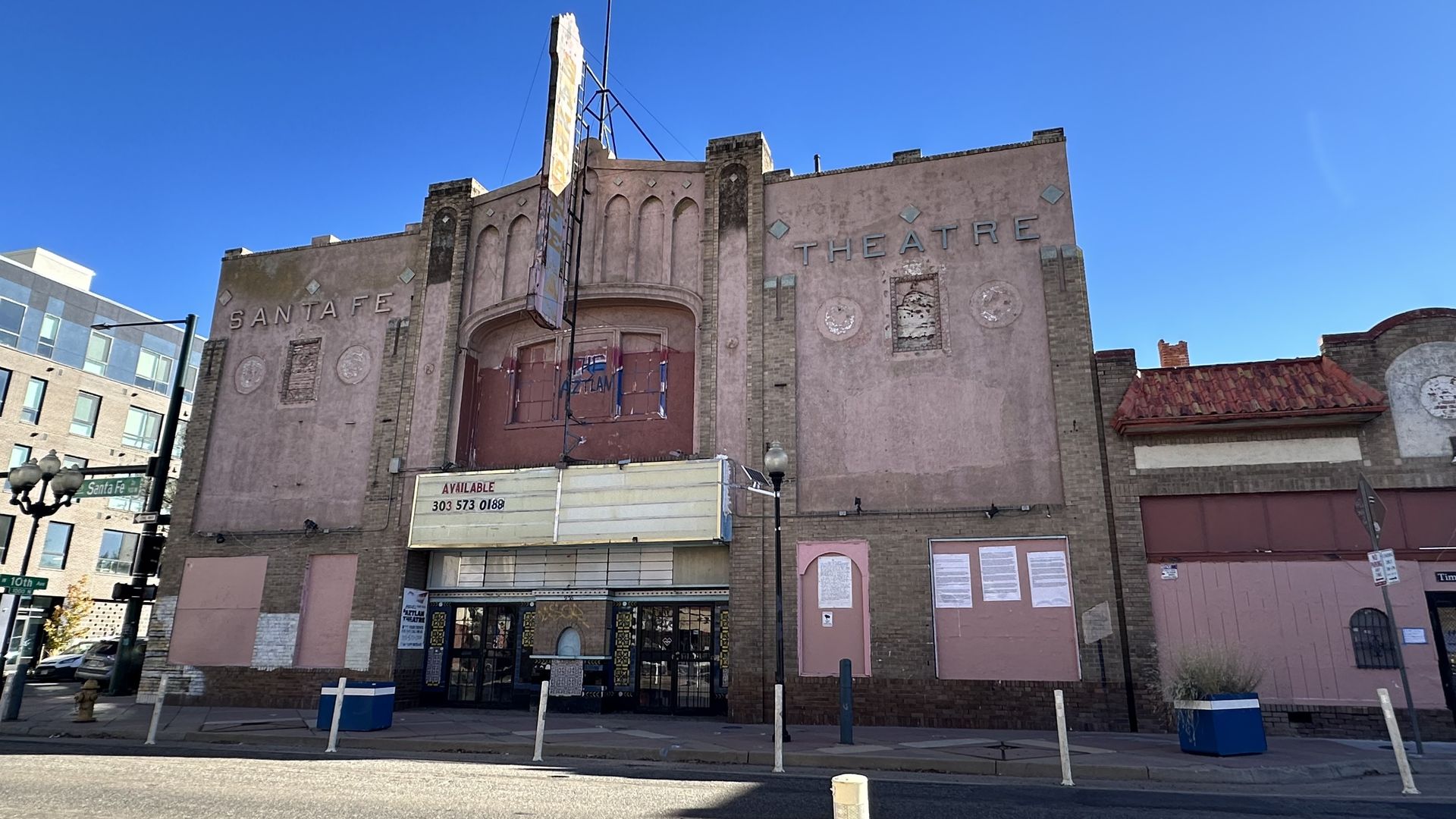 Old beige-pink Santa Fe Theatre façade with faded decorative panels, large "SANTA FE THEATRE" letters, a tall vertical sign, and a weathered brick entrance beneath a blue sky.