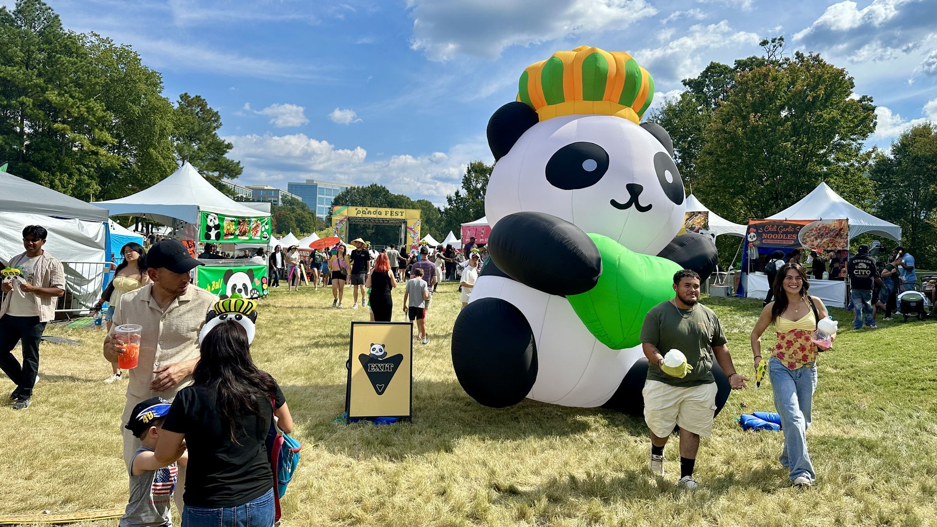Panda Fest Charlotte. Outdoor festival with an inflatable panda wearing a crown and holding a green leaf, booths and people walking on grass under a bright blue sky with clouds.