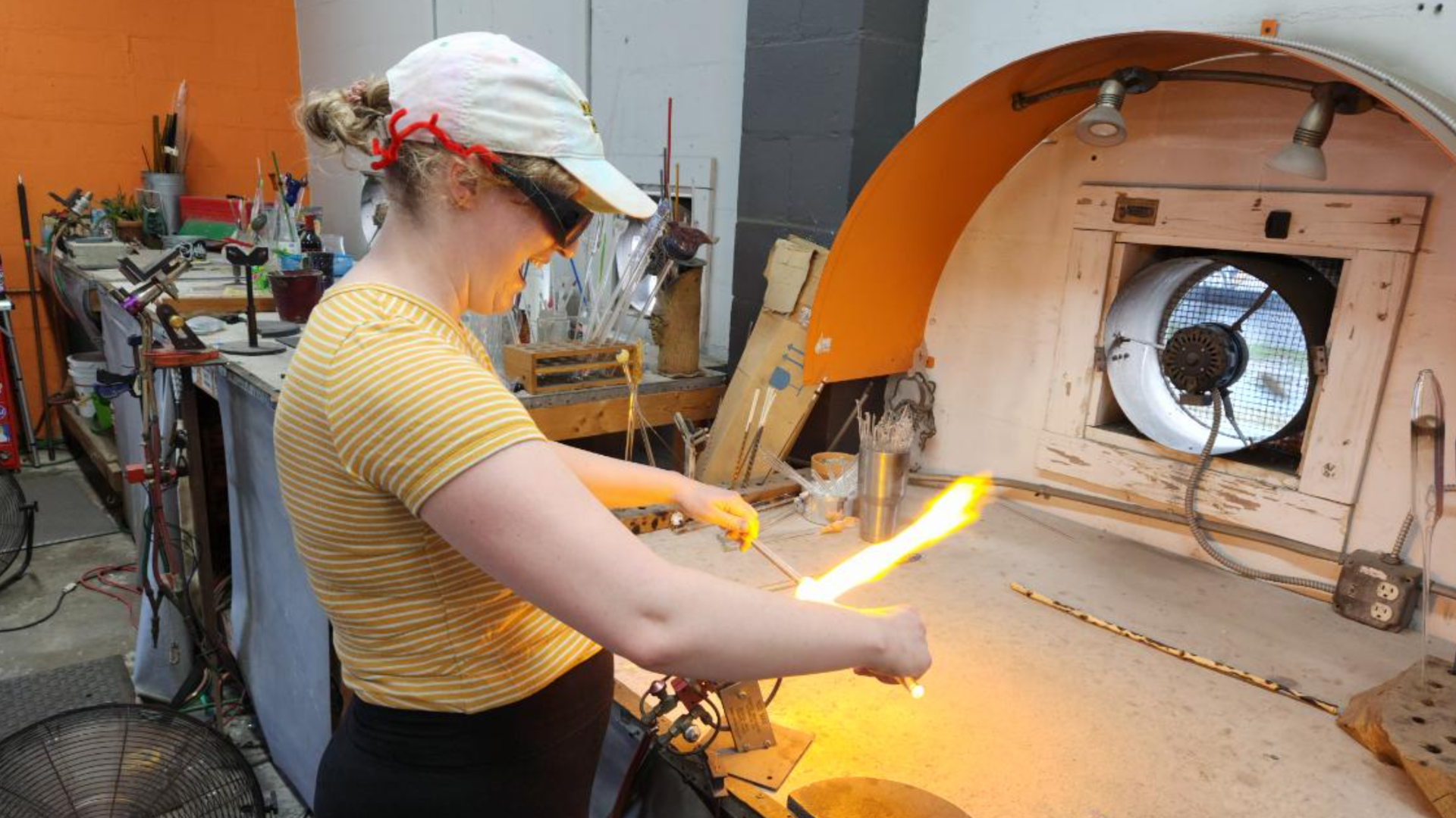 A woman in a yellow top and tie-dye baseball gap melts tubes of glass over an open flame.