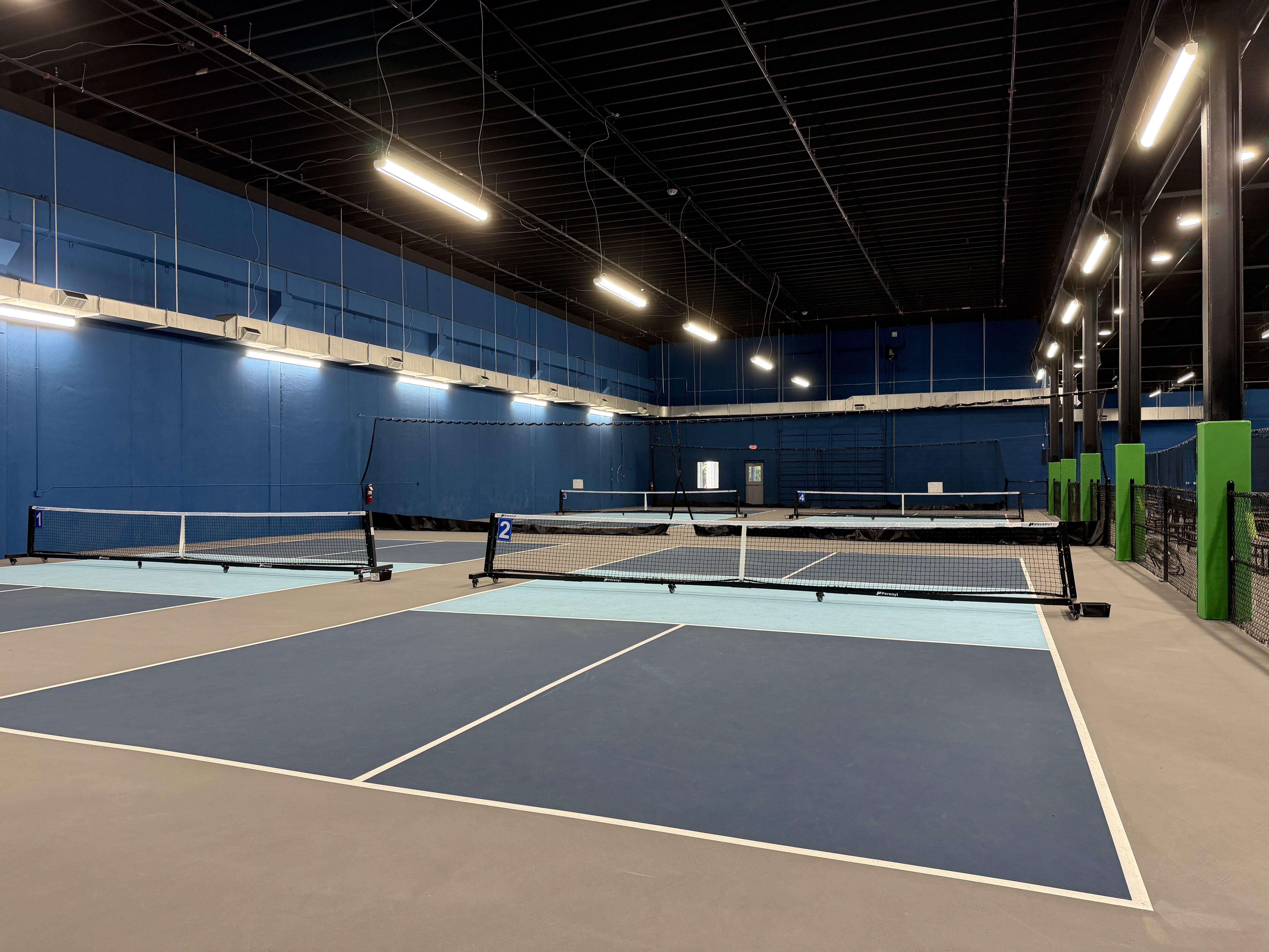 Indoor tennis facility with several blue courts, white boundary lines, and nets. Bright overhead lights illuminate the dark ceiling and blue walls, with black metal columns on the right.