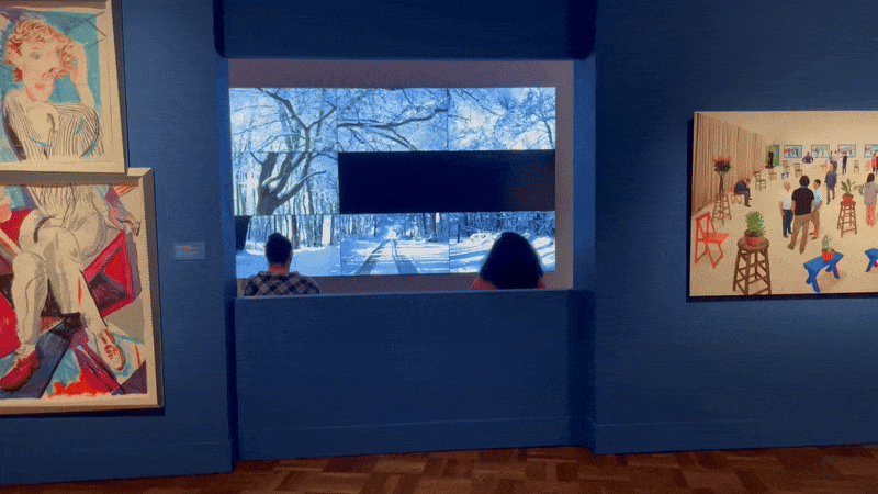 Art gallery with deep blue walls and framed paintings on either side of a large central video screen showing a snowy forest road. Two people sit on a bench facing the winter scene, wooden floor below.