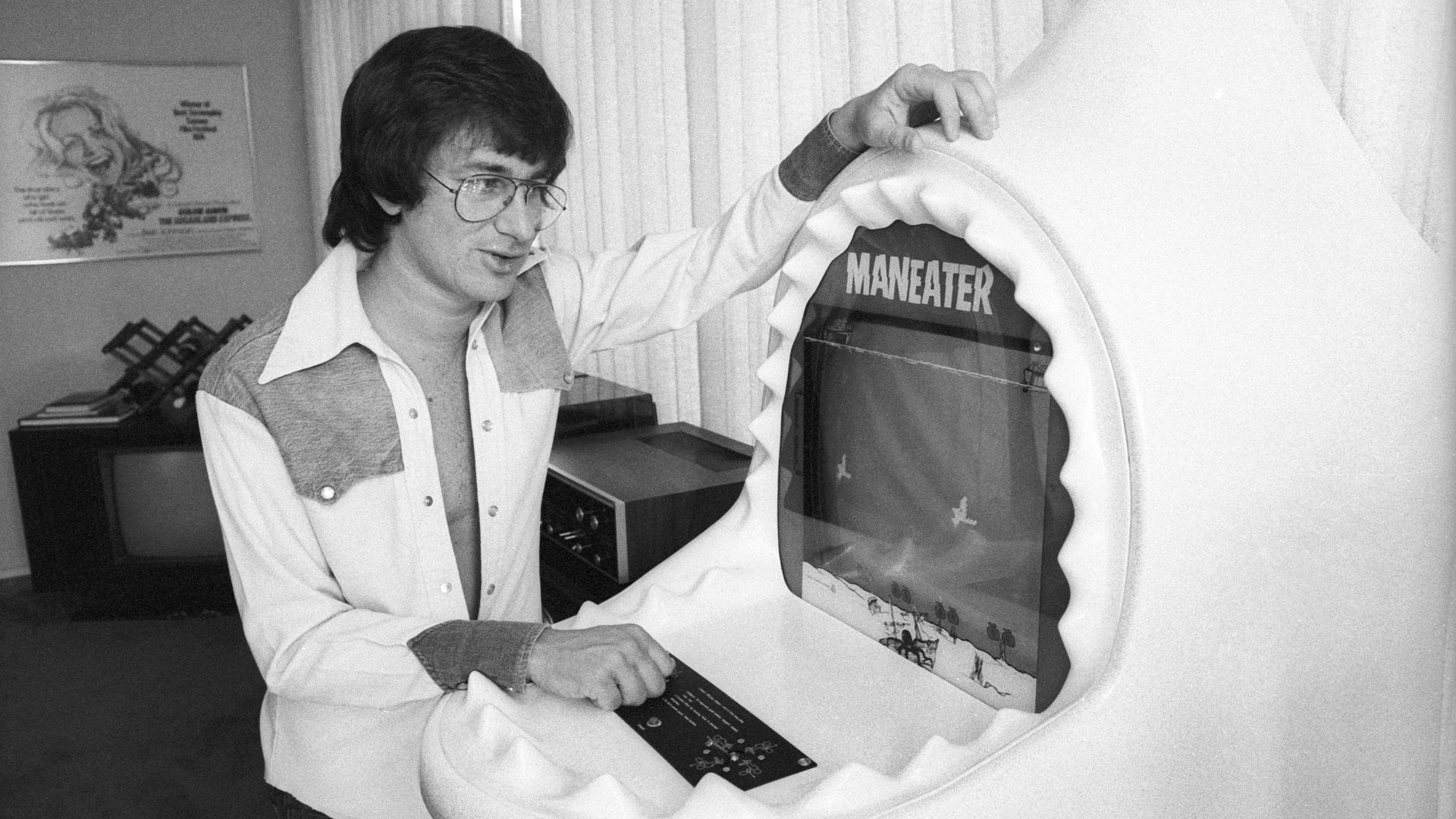 Black and white photo from the 1970s of a man standing next to an arcade game cabinet that is shaped like a shark with its jaws open
