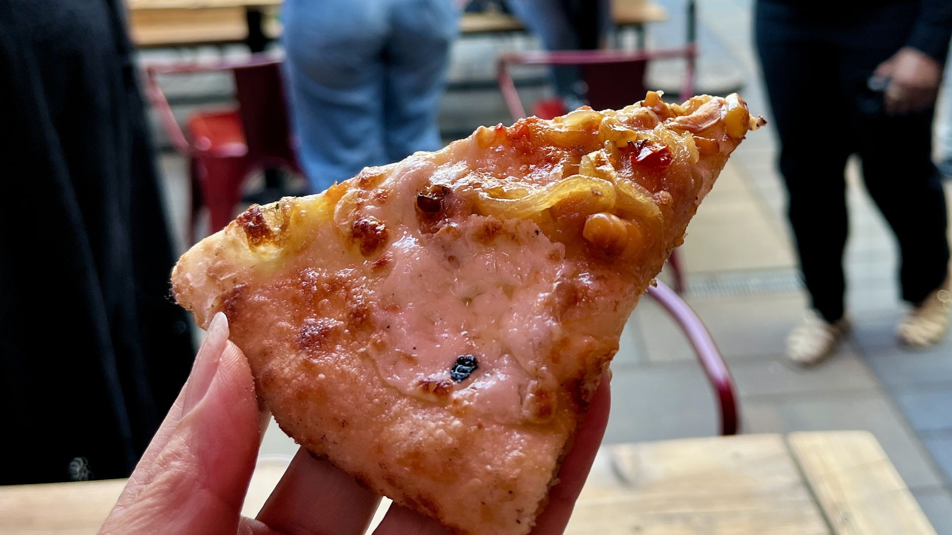 Close-up of a hand holding a slice of pizza with cheese and caramelized onions, with blurred people and tables in the background in an outdoor setting.