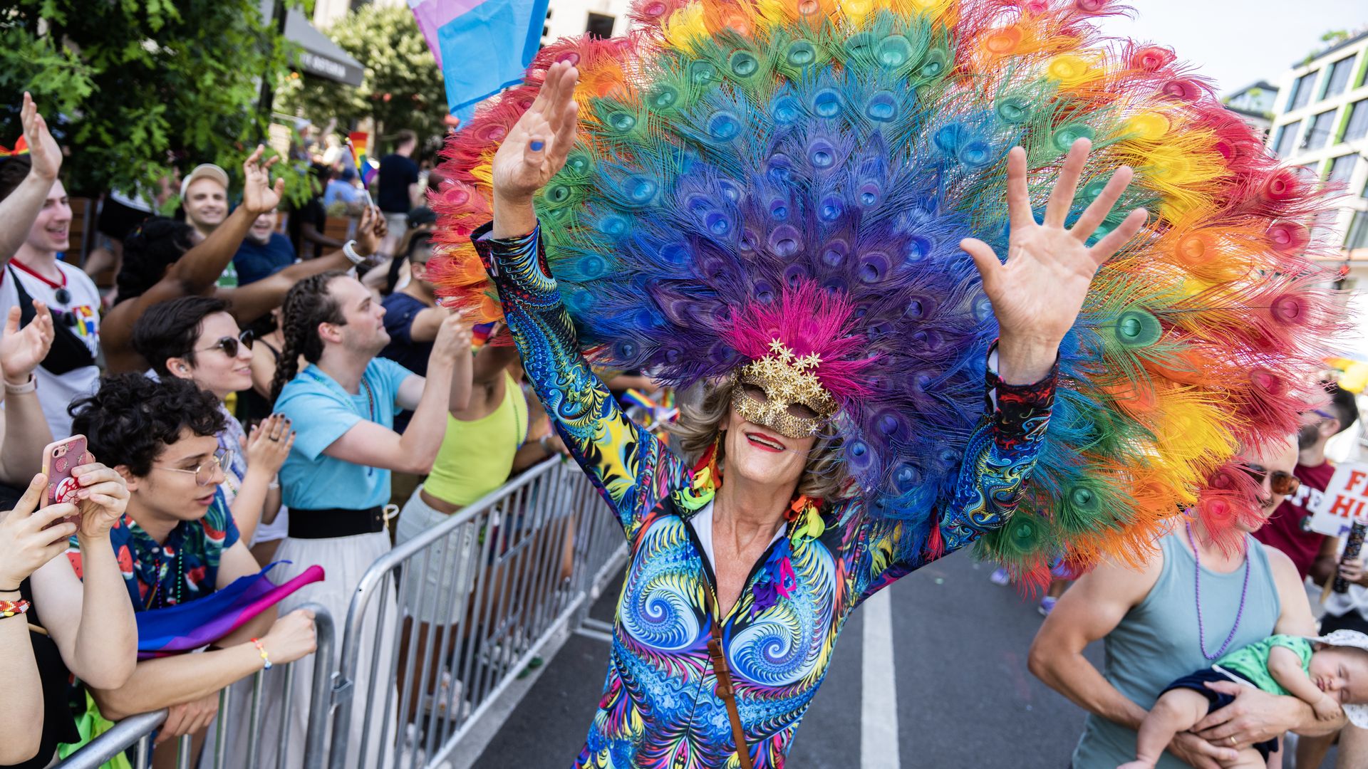A person dressed in a colorful suit and rainbow feather headdress greets a crowd at Capital Pride