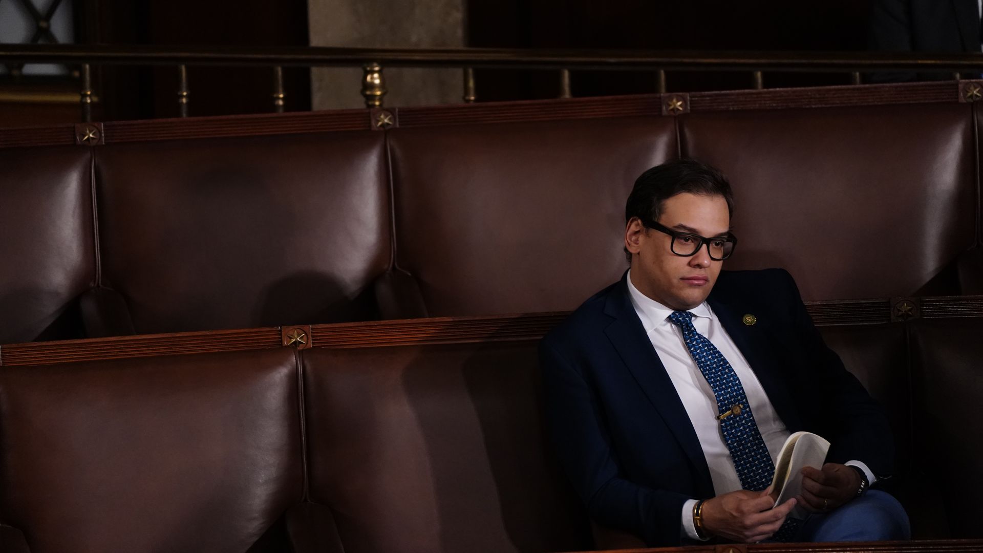Rep. George Santos, wearing a dark blue suit, white shirt, blue tie and thick-rimmed glasses, sits alone in the House chamber.