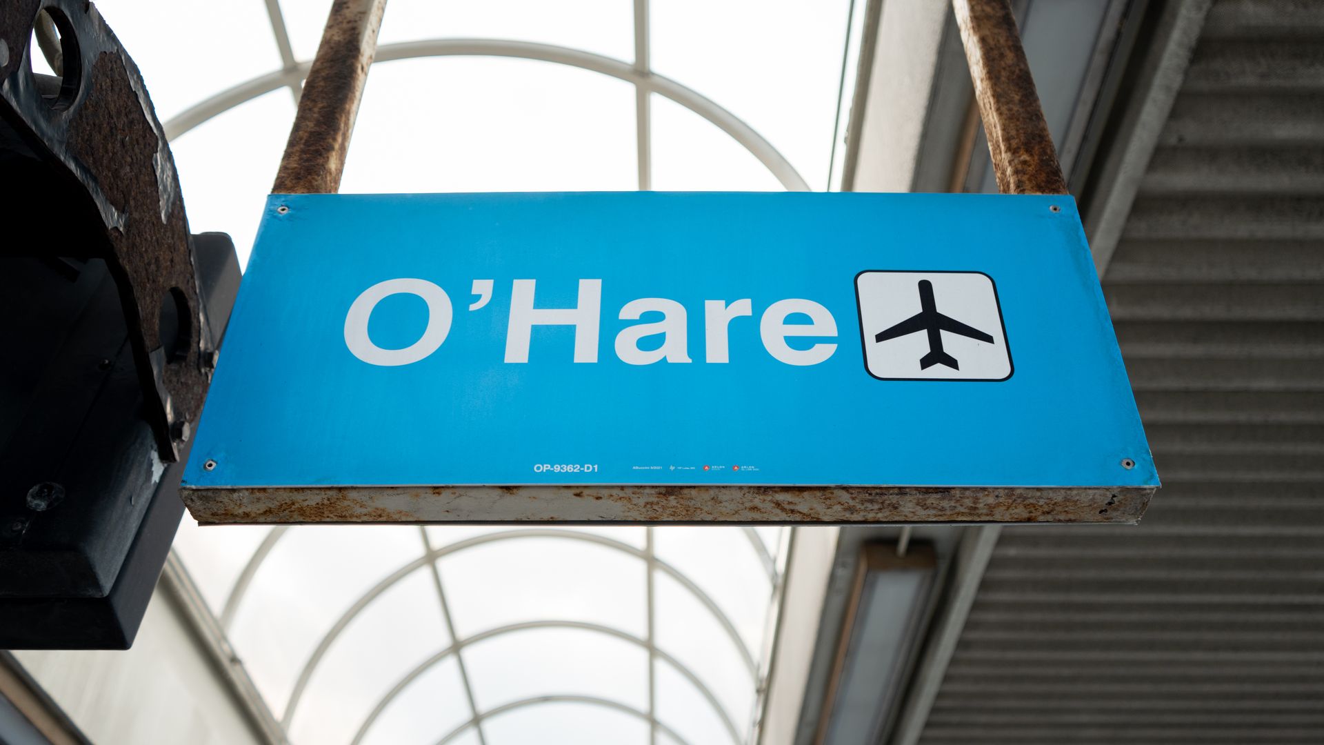 Blue O'Hare sign with a white airplane icon hangs from rusted metal beams in a covered airport corridor.