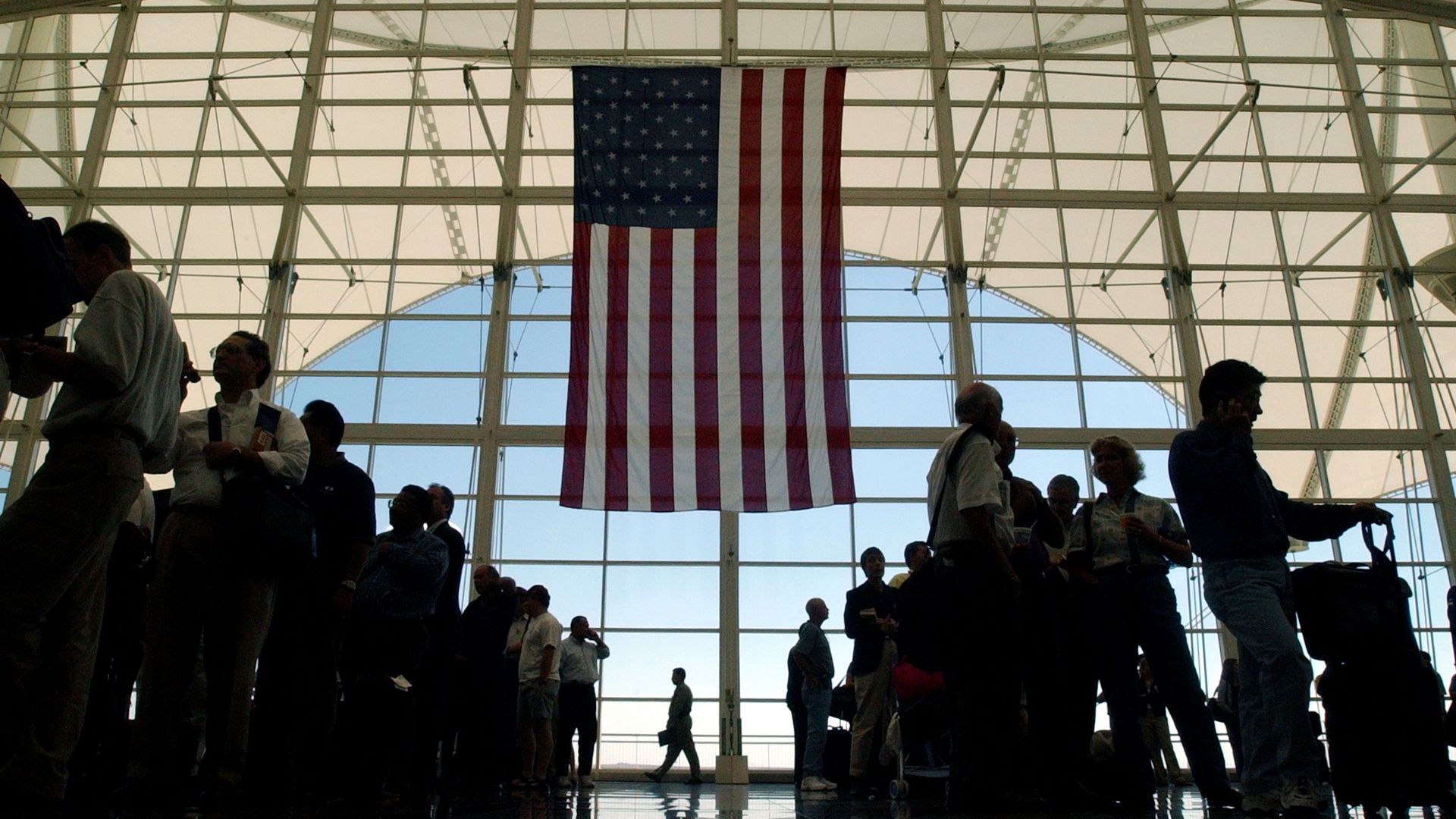 Inside a large glass atrium, silhouettes of people stand along a balcony beneath a hanging U.S. flag. Daylight filters through grid-like windows, highlighting the red, white, and blue stripes.