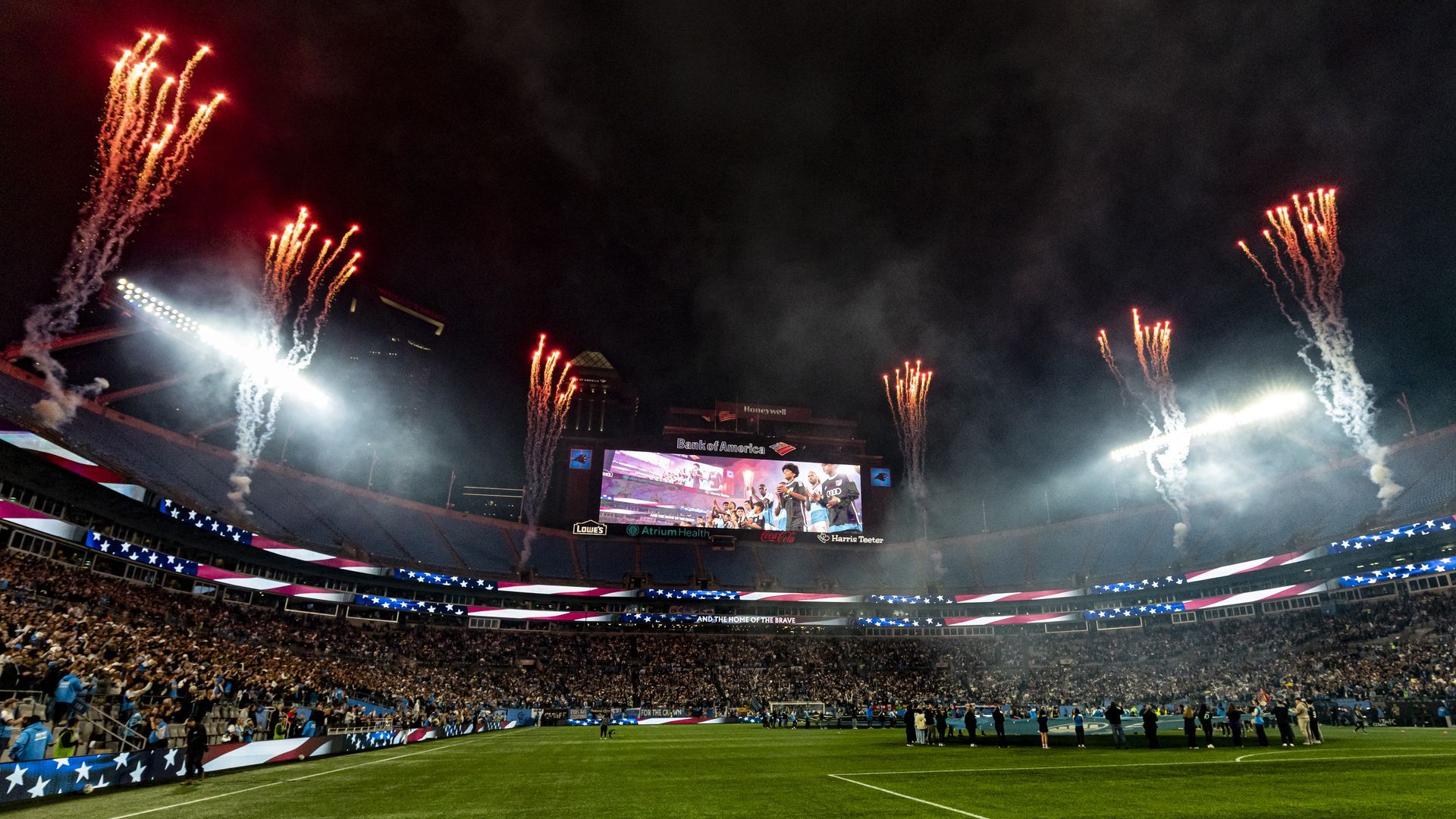 Nighttime stadium packed with a crowd, fireworks in the sky, and red, white, and blue lights and banners with stars displayed, celebrating a patriotic event.