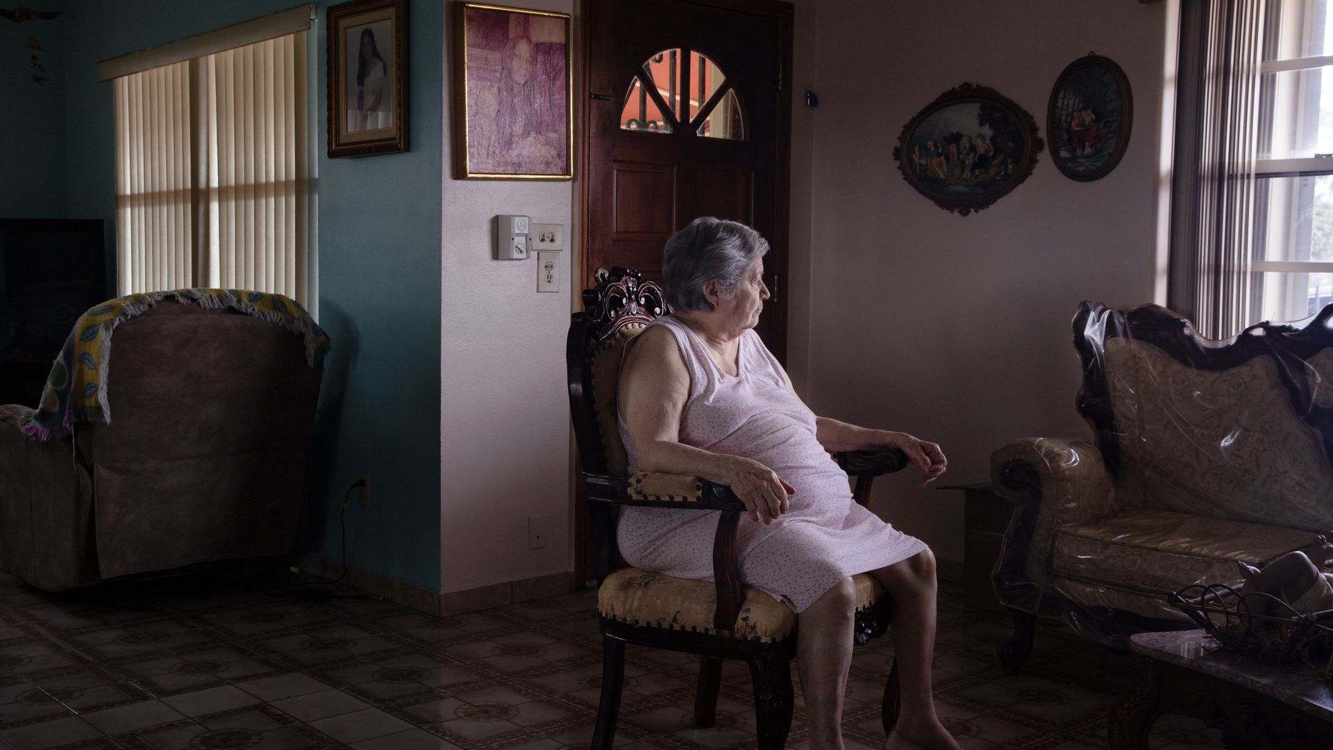 Elderly woman in a light dress sitting on an ornate wooden chair in a dimly lit living room, looking out a window with vertical blinds during daytime.