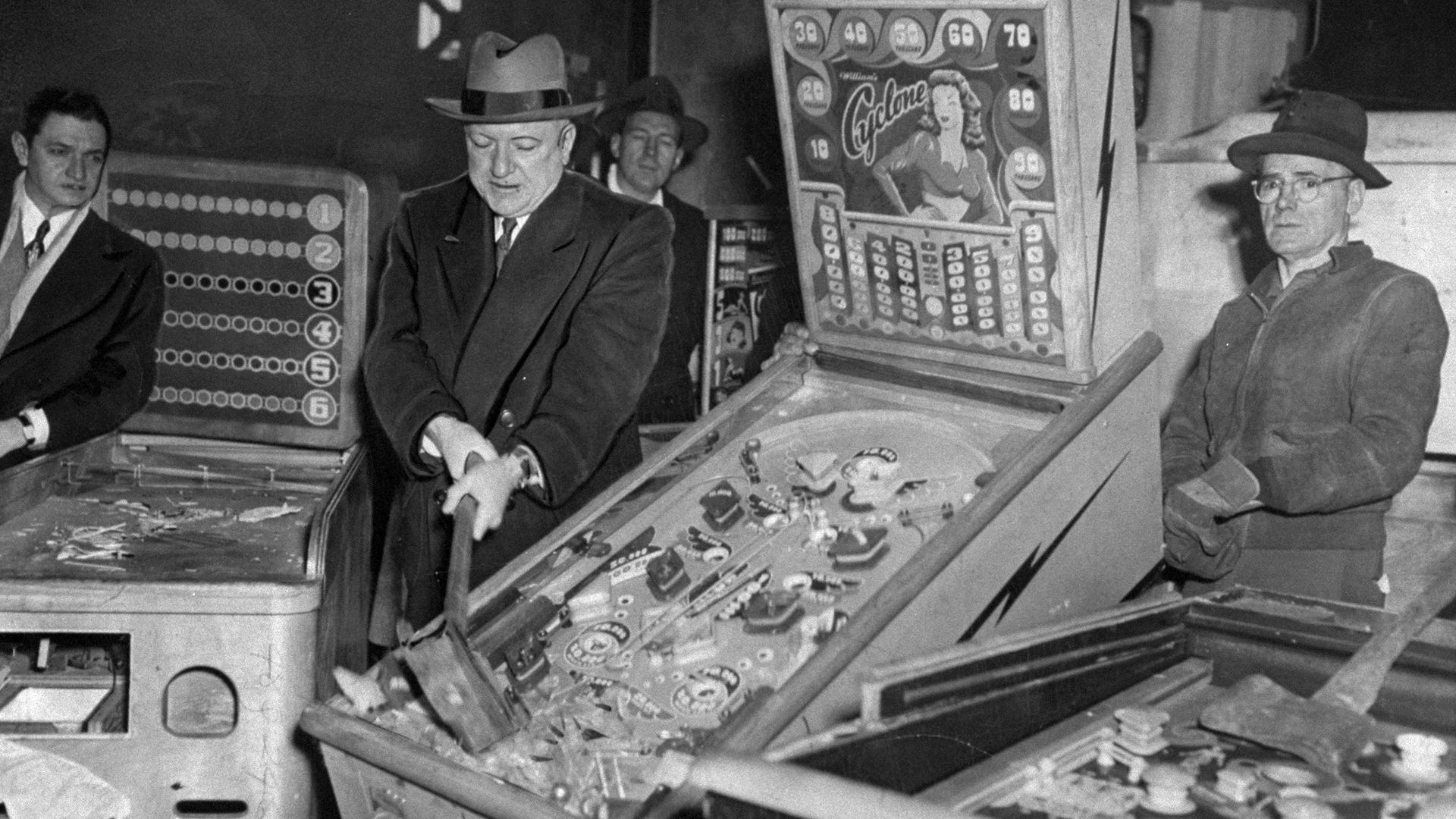 A New York City police commissioner takes a sledgehammer to a pinball machine's glass casing as others look on.
