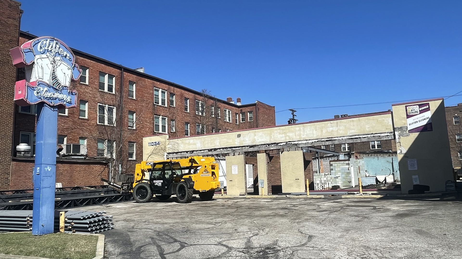 Clear blue sky above a red brick building with a yellow construction vehicle in a yard. Foreground shows stacked metal pipes and a tall blue pole supporting a pink and blue neon sign.
