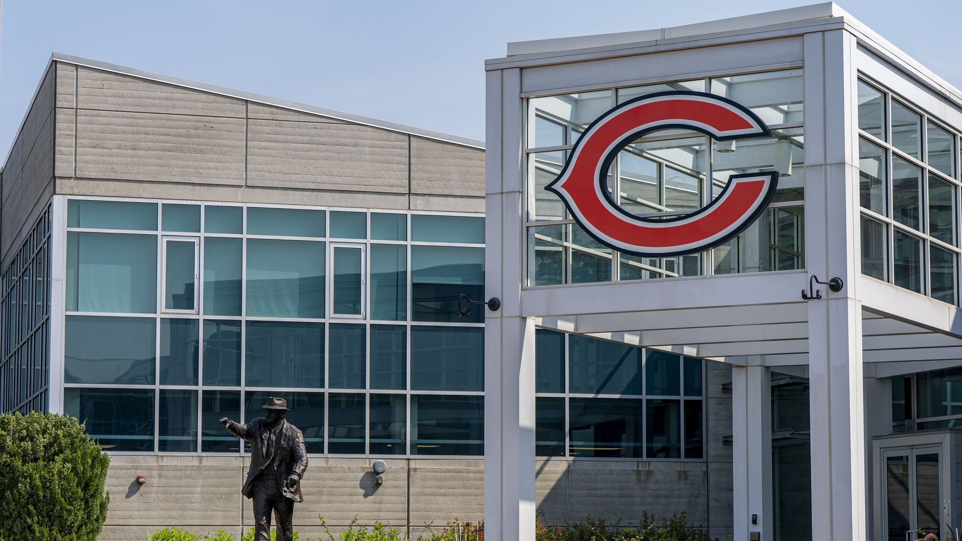 Modern glass-front building with a large red C sign, statue of George S. Halas in front, and trimmed landscaping under a clear blue sky.