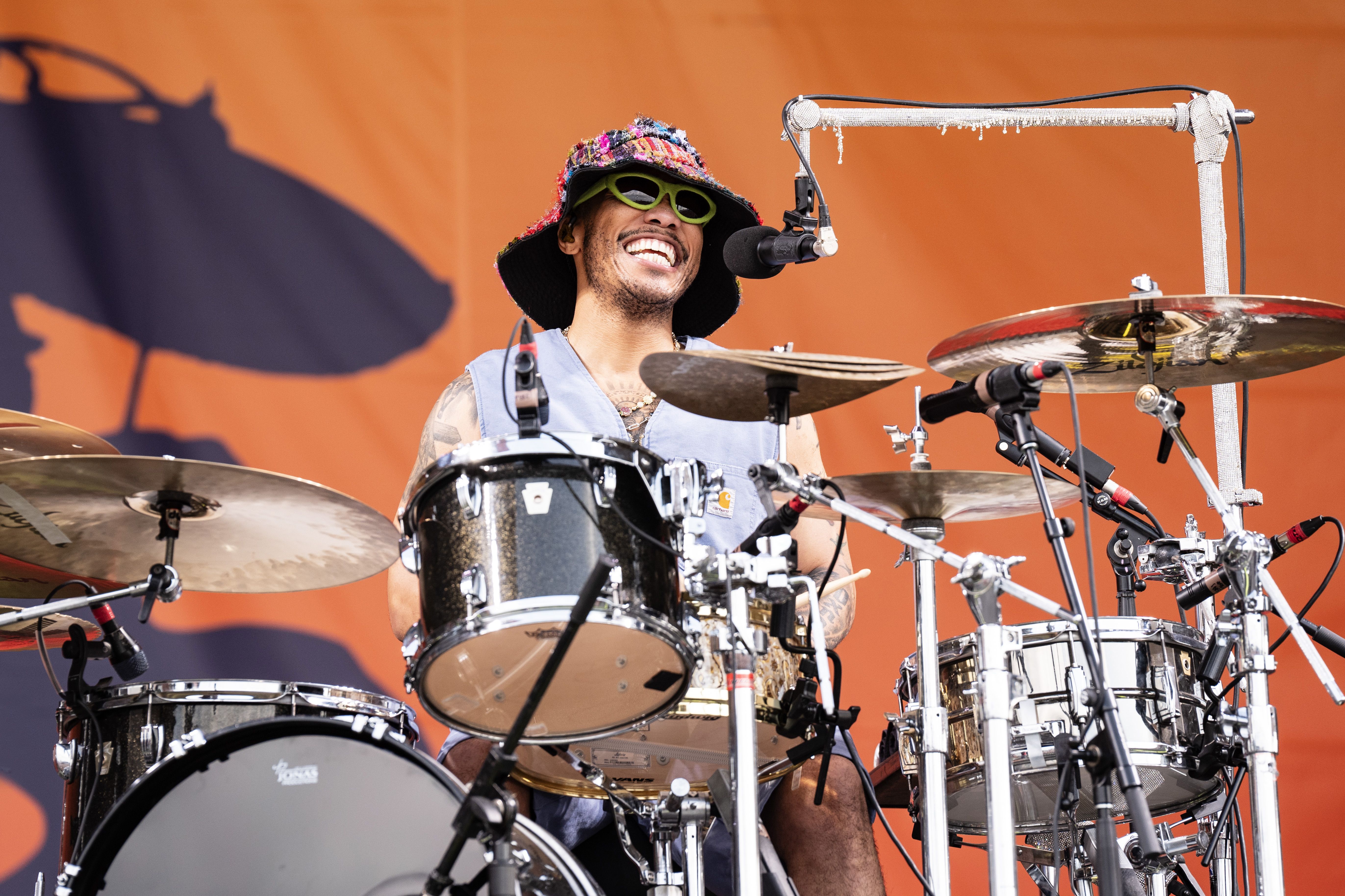 Anderson.Paak smiles at the microphone while drumming.