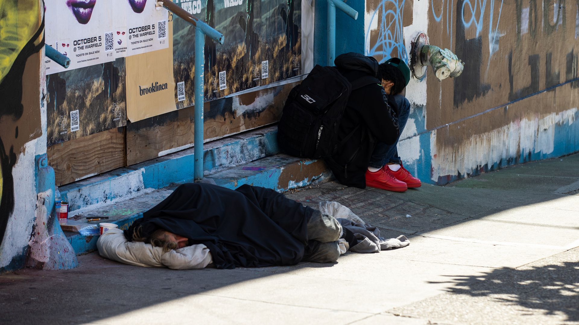 Photo of two people crouched against a wall trying to sleep on the sidewalk