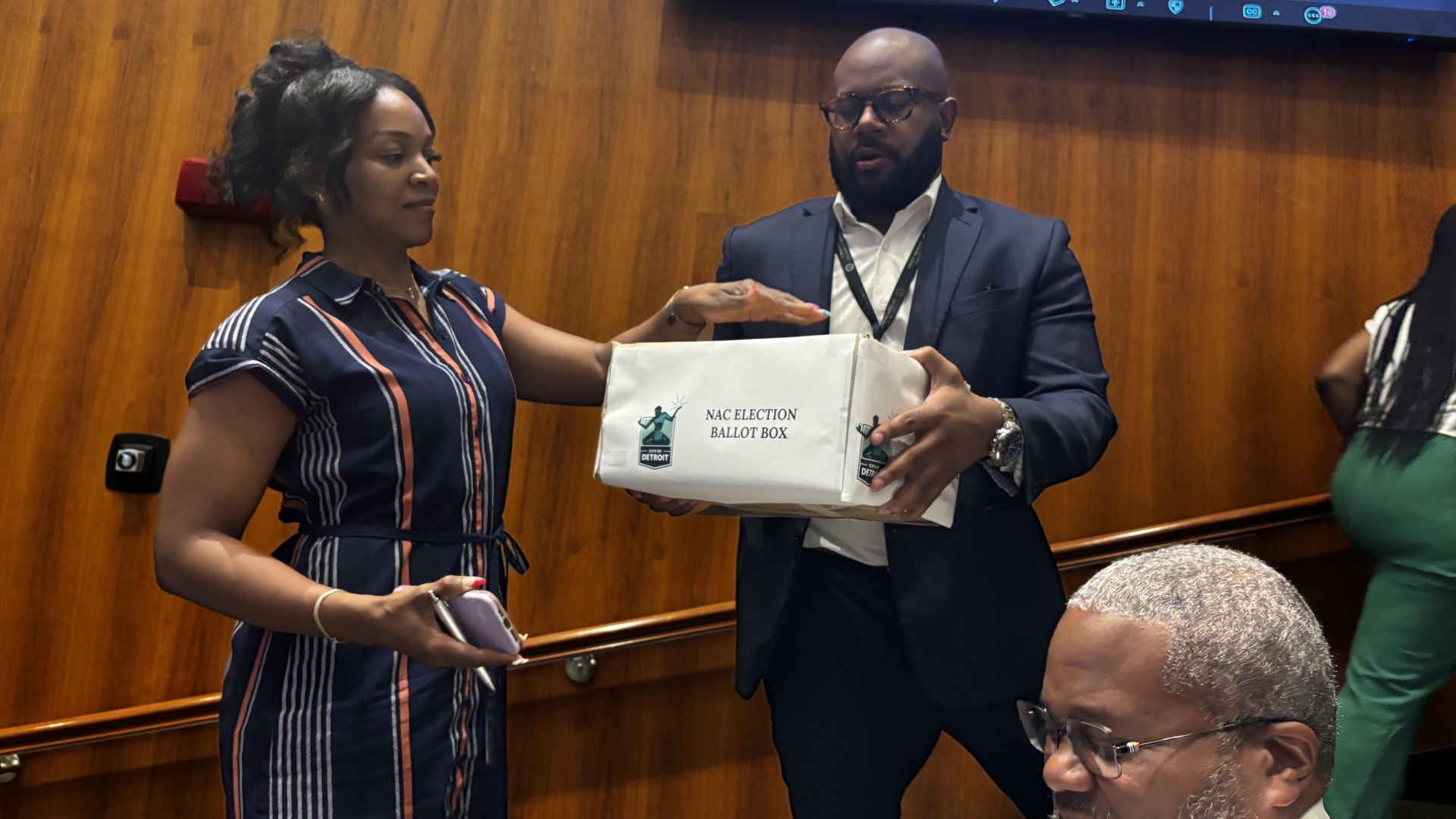 Two professionals exchange a white ballot box bag in a wood-paneled room; a man in a suit and glasses receives it from a woman as a projector screen glows blue above.