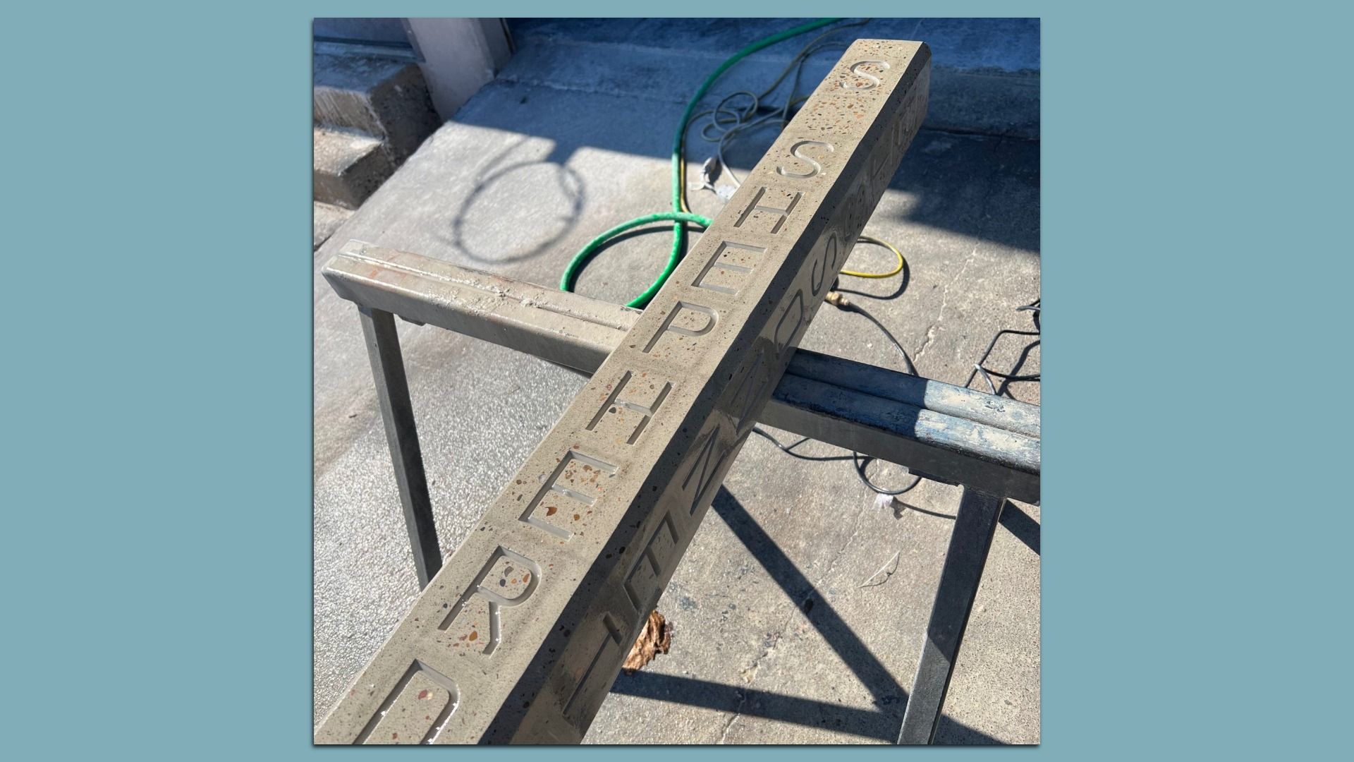 Concrete block with the words "SHEPHERD" carved into it, resting on a metal sawhorse in an outdoor workspace with a green hose and concrete floor visible around it.