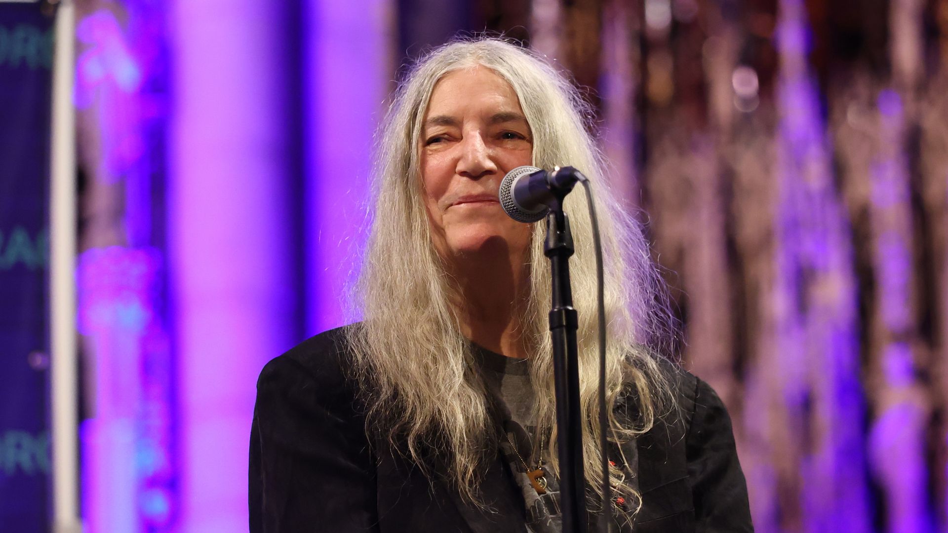 Portrait of an older woman with long gray hair singing into a microphone on a stage, purple lighting in the background.