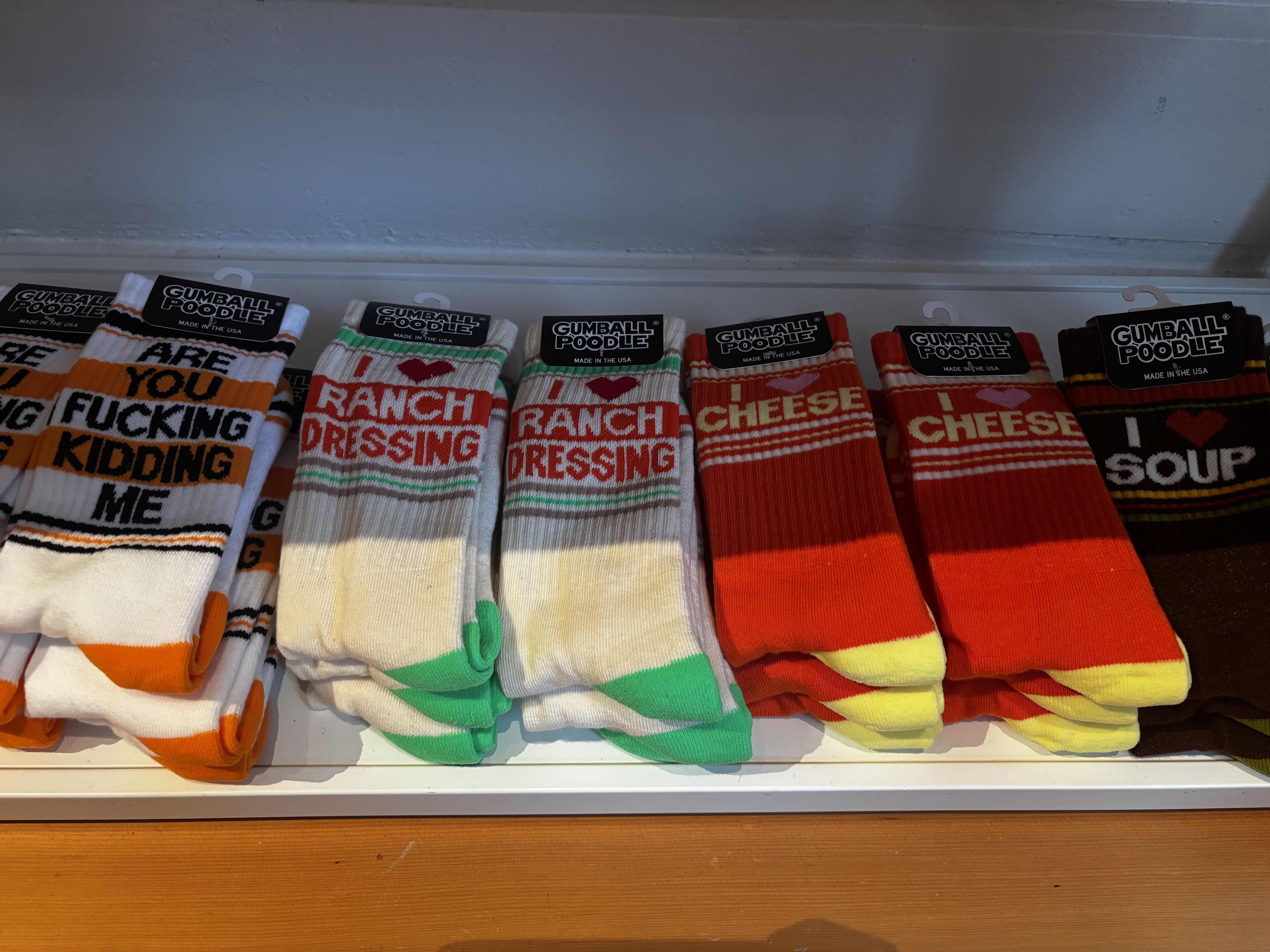Six pairs of colorful socks on a shelf with text: white-orange "ARE YOU FUCKING KIDDING ME," white-green "I โฅ RANCH DRESSING," red-yellow "I โฅ CHEESE," and brown-yellow "I โฅ SOUP."