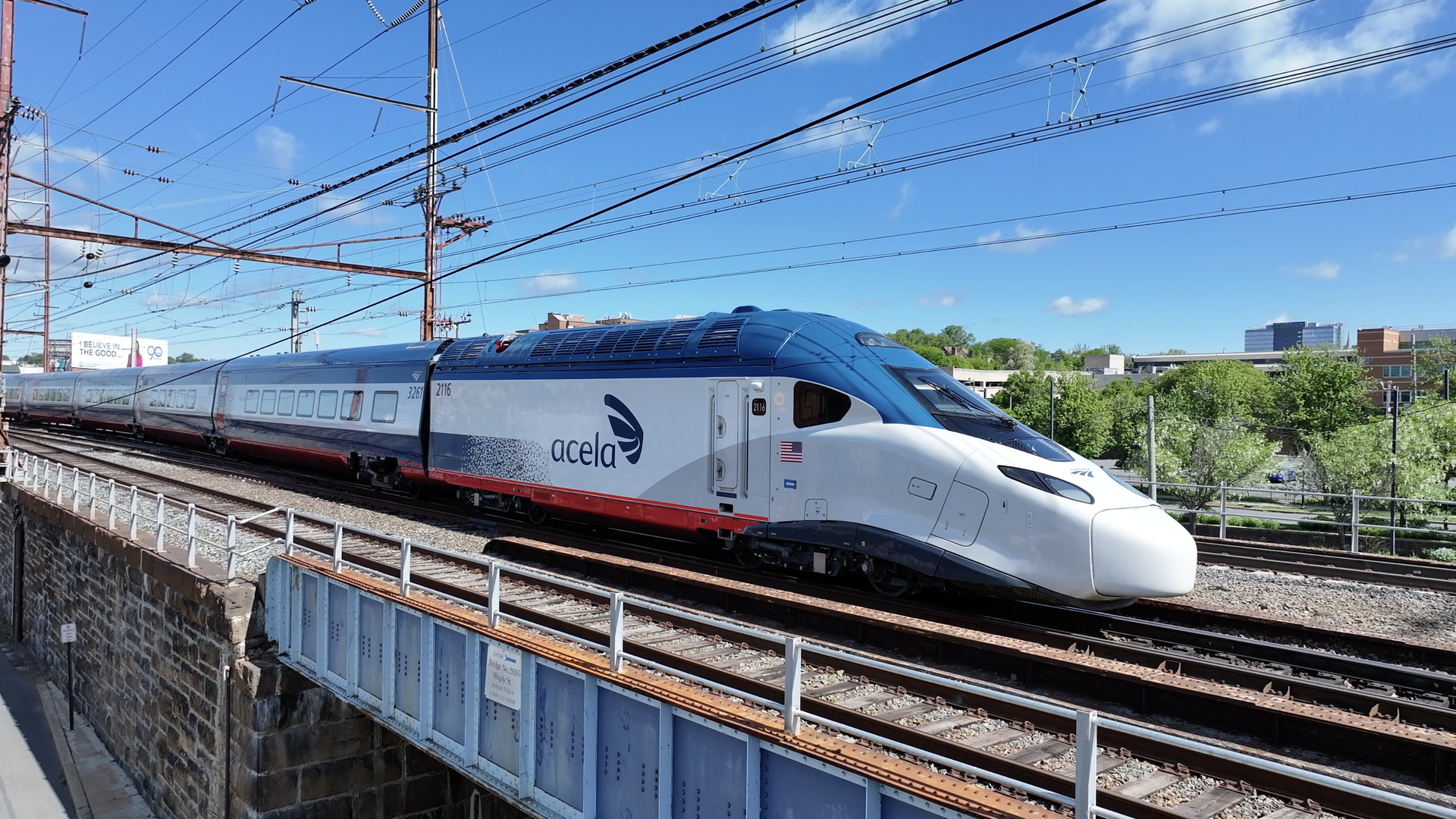 Amtrak's new NextGen Acela high-speed train traveling on elevated tracks under blue sky with some buildings and green trees in background.