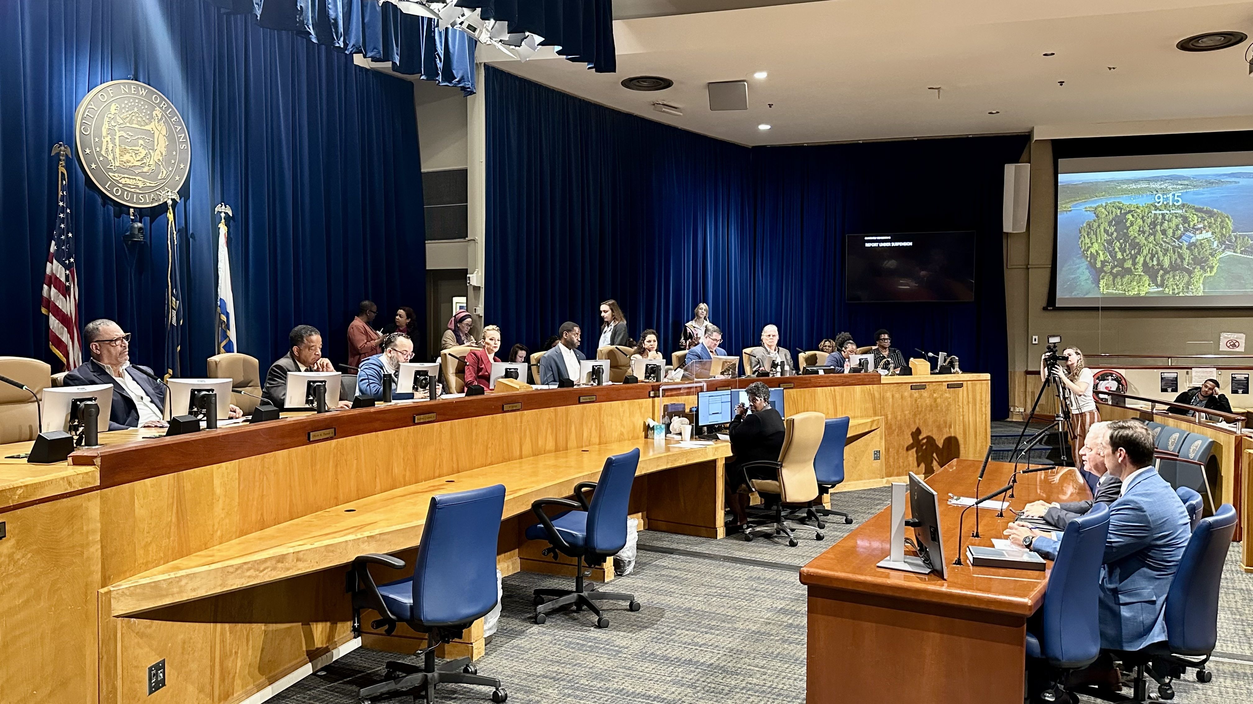 City council meeting in New Orleans chamber with members seated at a semicircular wooden desk, blue chairs, blue curtains, American and Louisiana flags, and screens showing an island and documents.