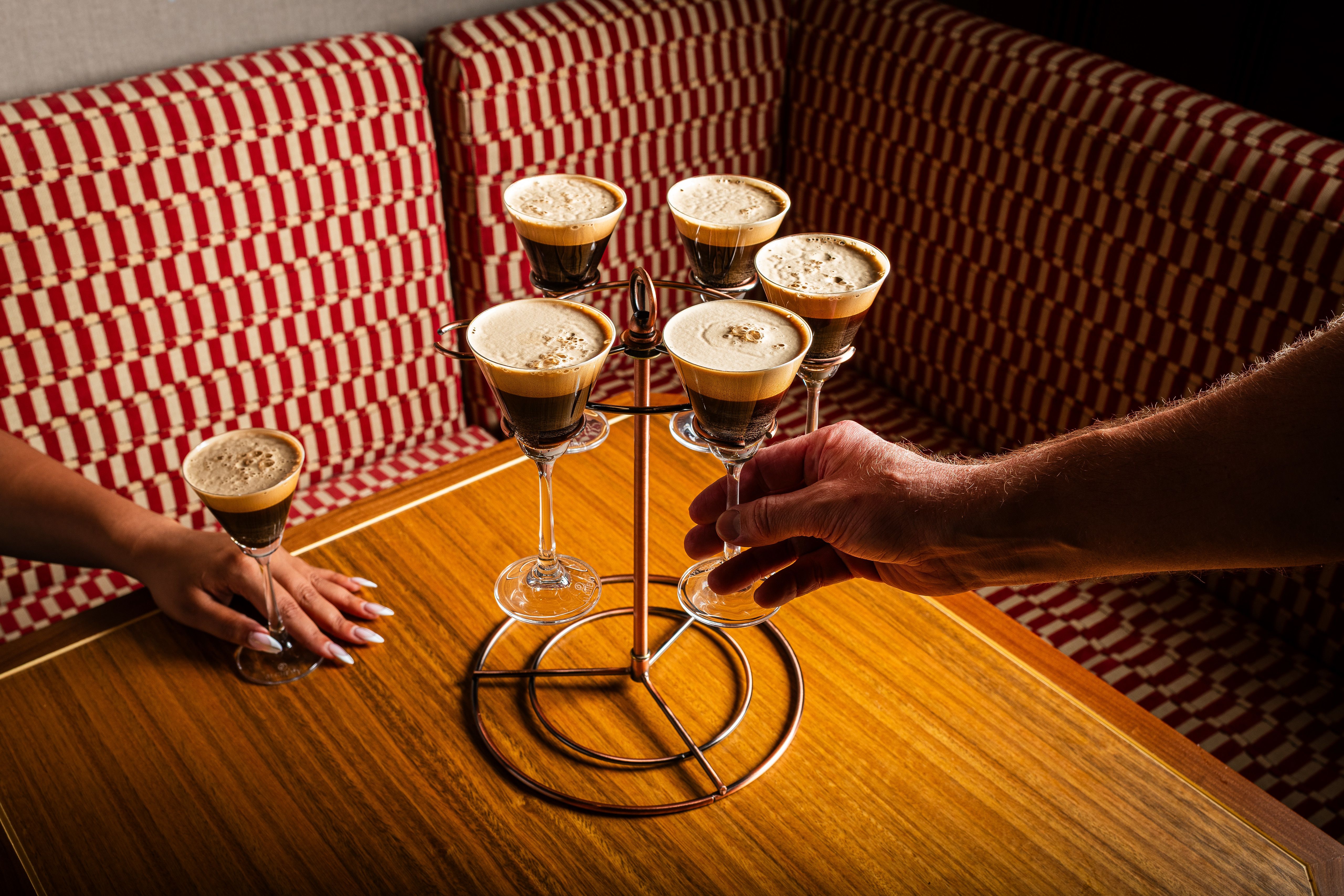 On a wooden table, a copper multi-tier stand holds several espresso cocktails in stemmed glasses with dark coffee layers and light foam; two hands steady the rack, red-and-white checkered upholstery in the background.
