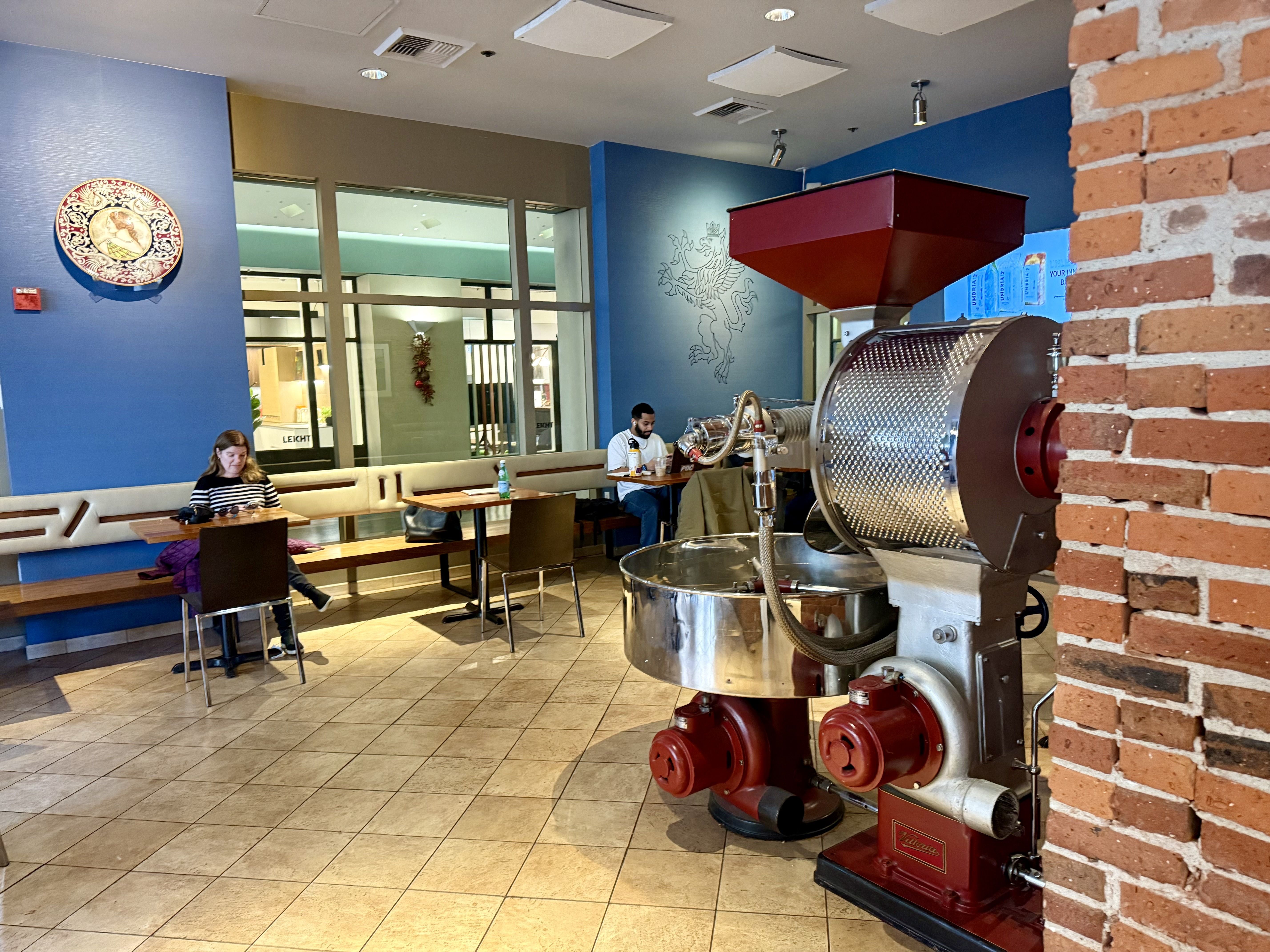 Interior of a coffee shop with blue walls, a large red and silver coffee roasting machine, tiled floor, wooden tables, and two people sitting separately, one working on a laptop.