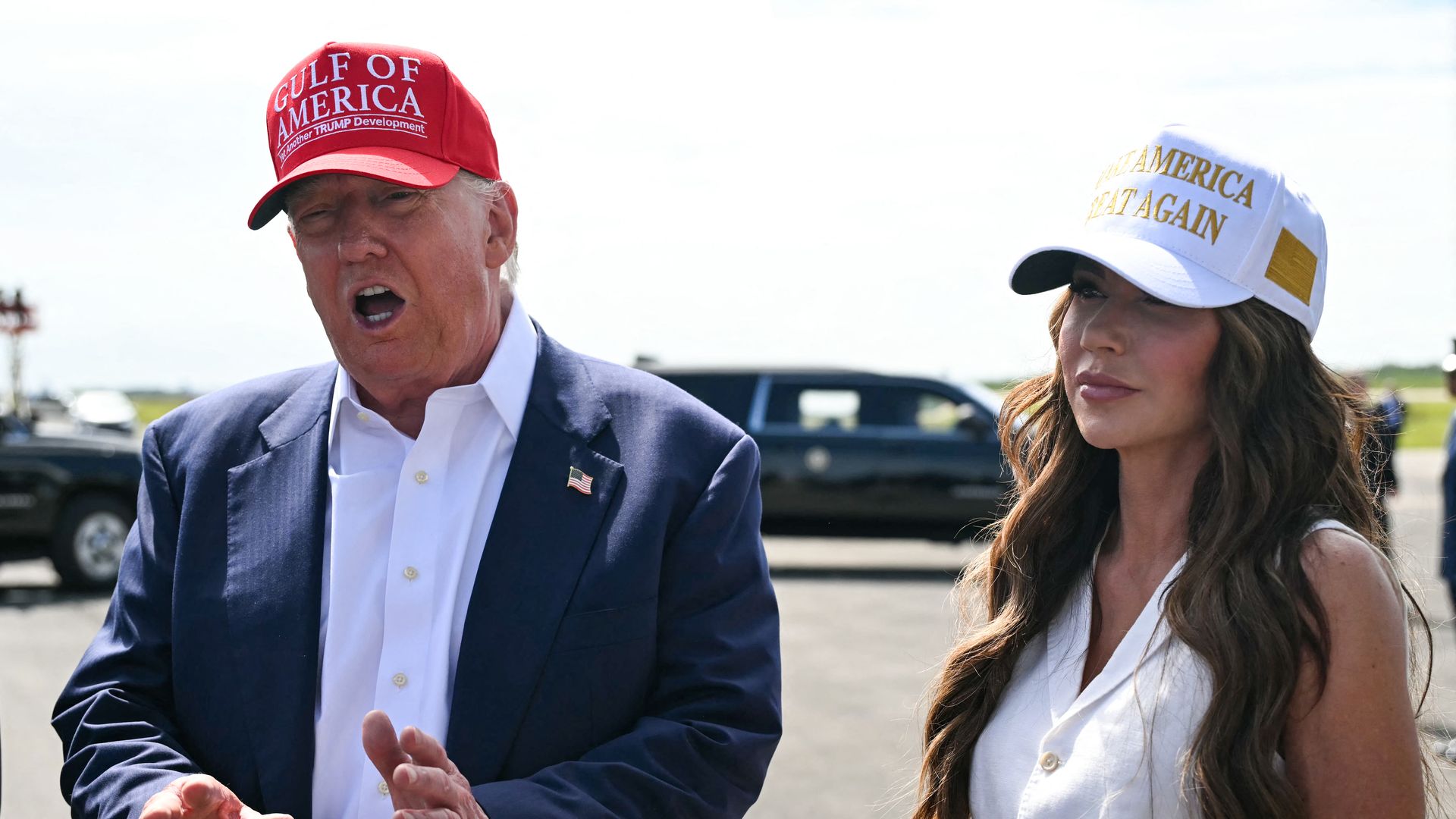 Man in navy suit and red hat, President Trump, and woman in white hat and white blouse, Homeland Security Secretary Kristi Noem, standing in an outside space.