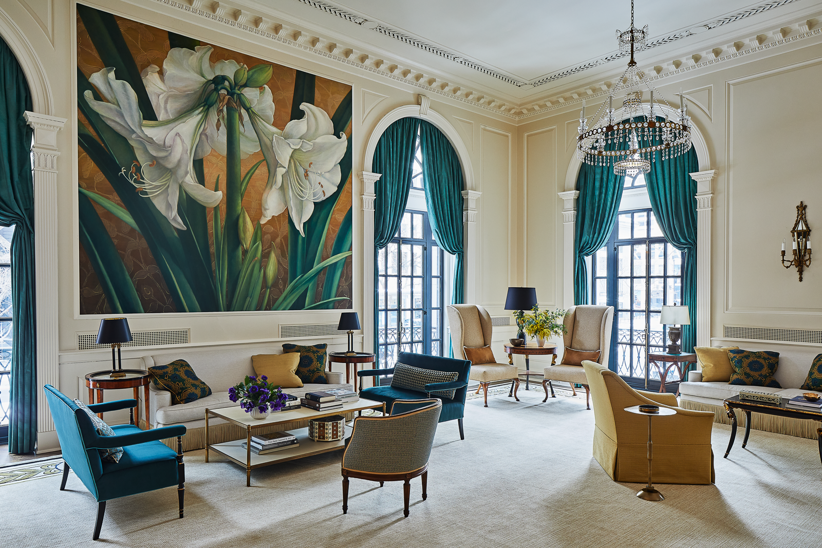 Luxurious living room featuring a large white lily mural, teal drapes, ornate molding, a crystal chandelier, and blue/beige seating around a central coffee table.