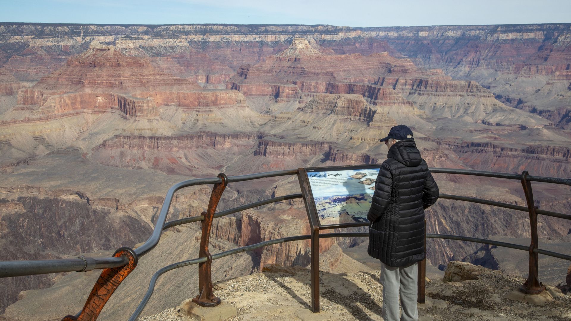 Tourist in a black puffer jacket stands at a rusted railing overlooking the layered red Grand Canyon, reading an information sign as blue sky stretches above.