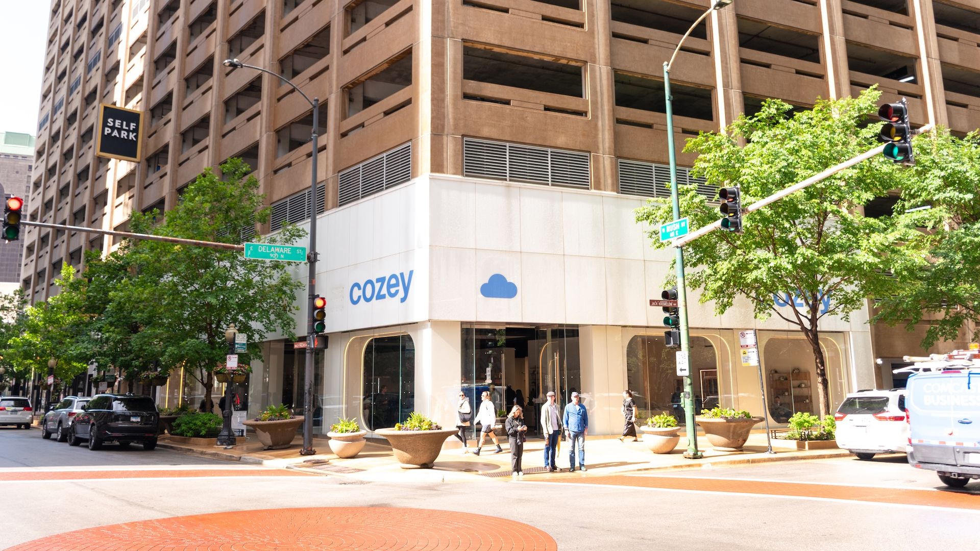 Urban street view showing a tall beige parking garage with a 'SELF PARK' sign, green trees, pedestrians on the sidewalk, traffic lights, and storefronts along the ground level.