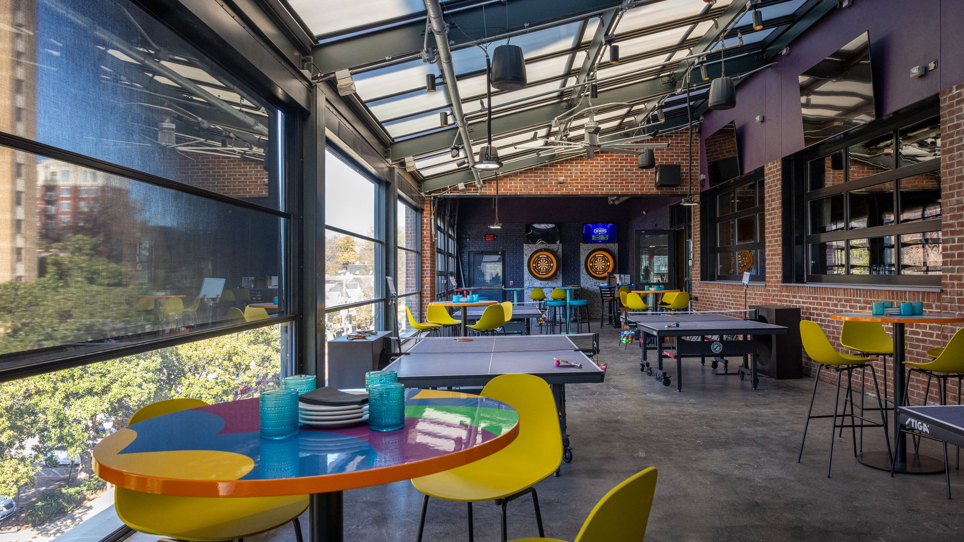 Indoor game room with yellow chairs and colorful tables, ping pong tables in the center, dartboards on brick walls, large windows, and a glass ceiling letting in natural light.
