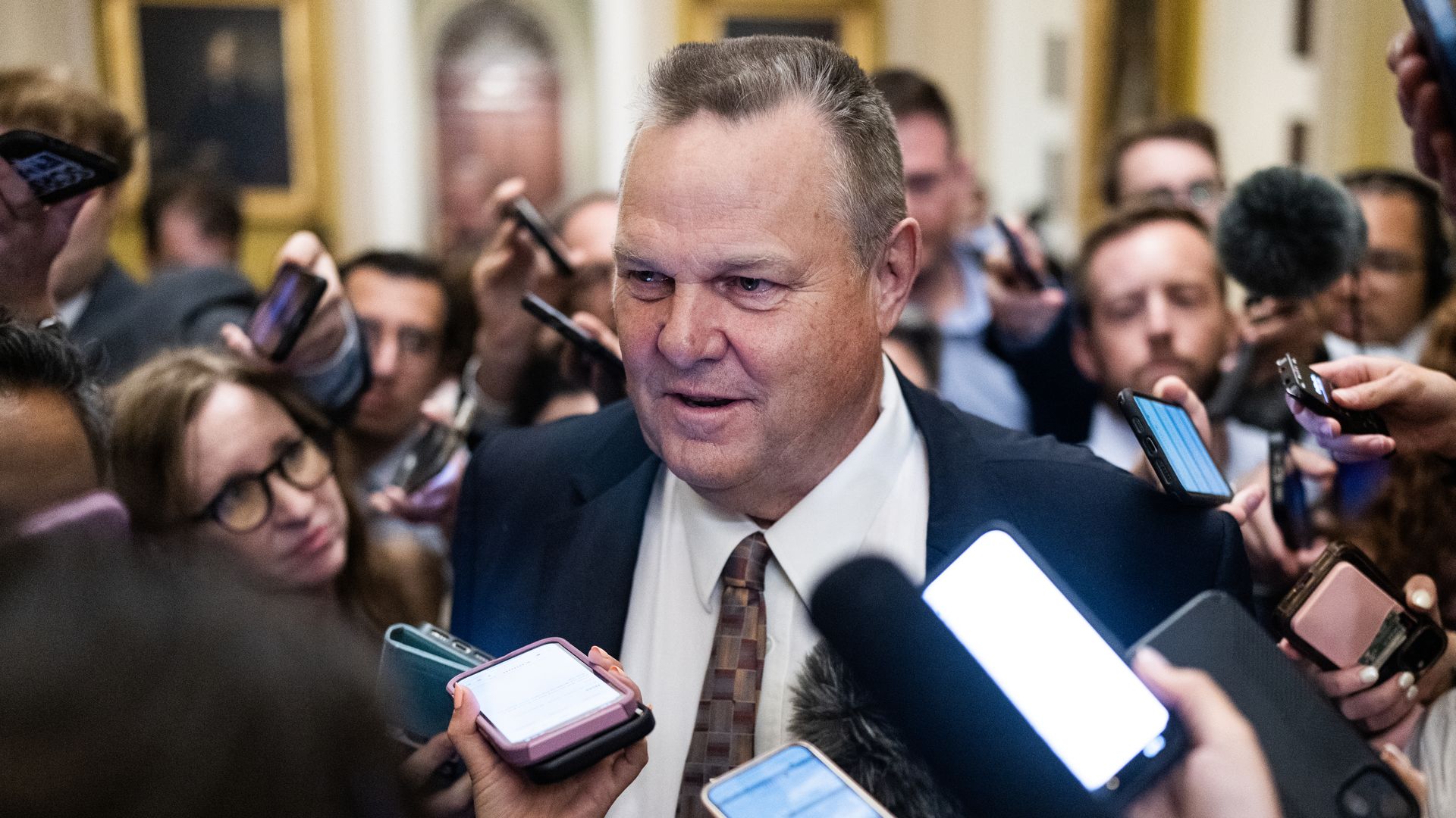 Sen. Jon Tester is surrounded by reporters with phones or recorders near his face. 