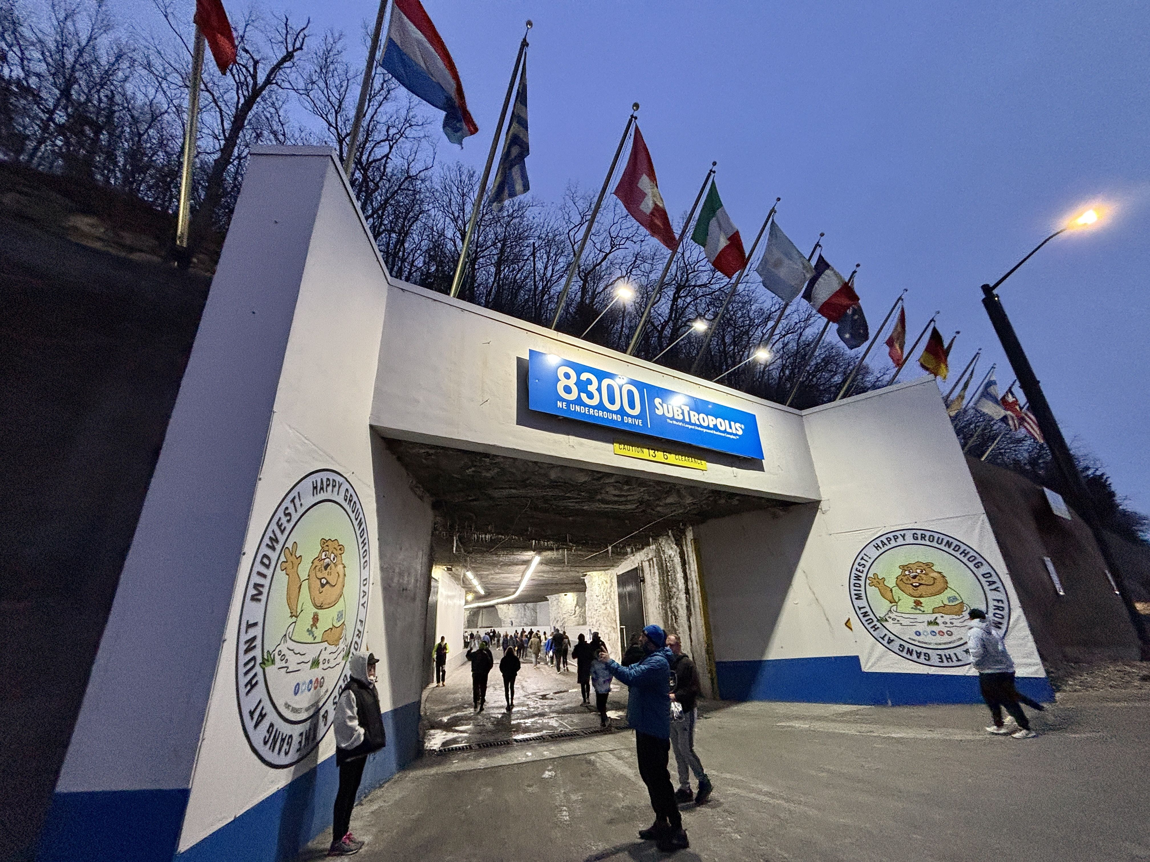 Tunnel entrance at 8300 NE Underground Drive labeled SubTropolis, with flags overhead. People stand outside and walk inside as dusk turns to night. Groundhog Day cartoon on walls.