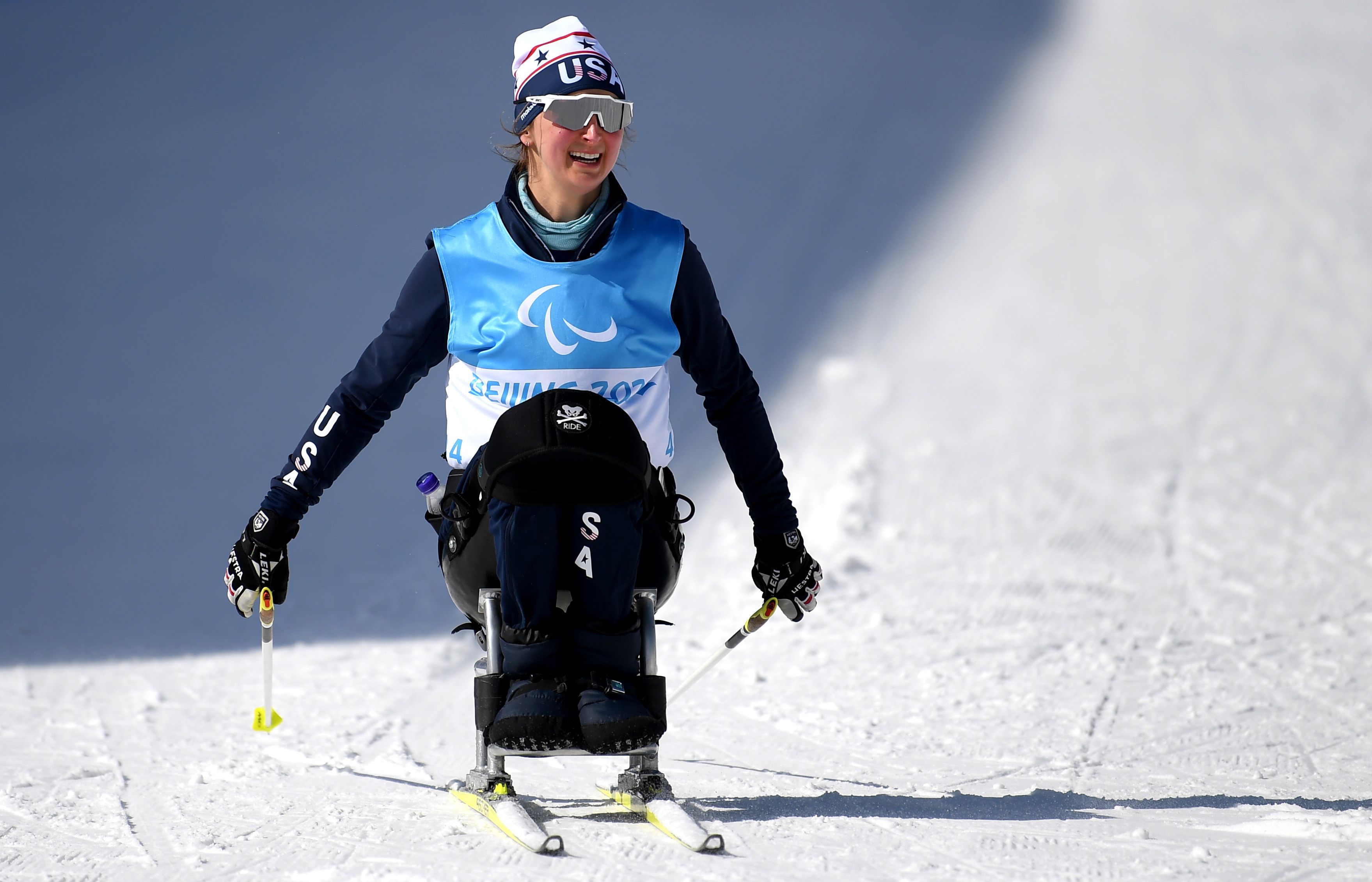 Smiling USA Paralympic skier Erin Martin in blue bib and white hat skiing on snow with dual ski poles, seated on a sit-ski during Beijing 2022 Winter Paralympics